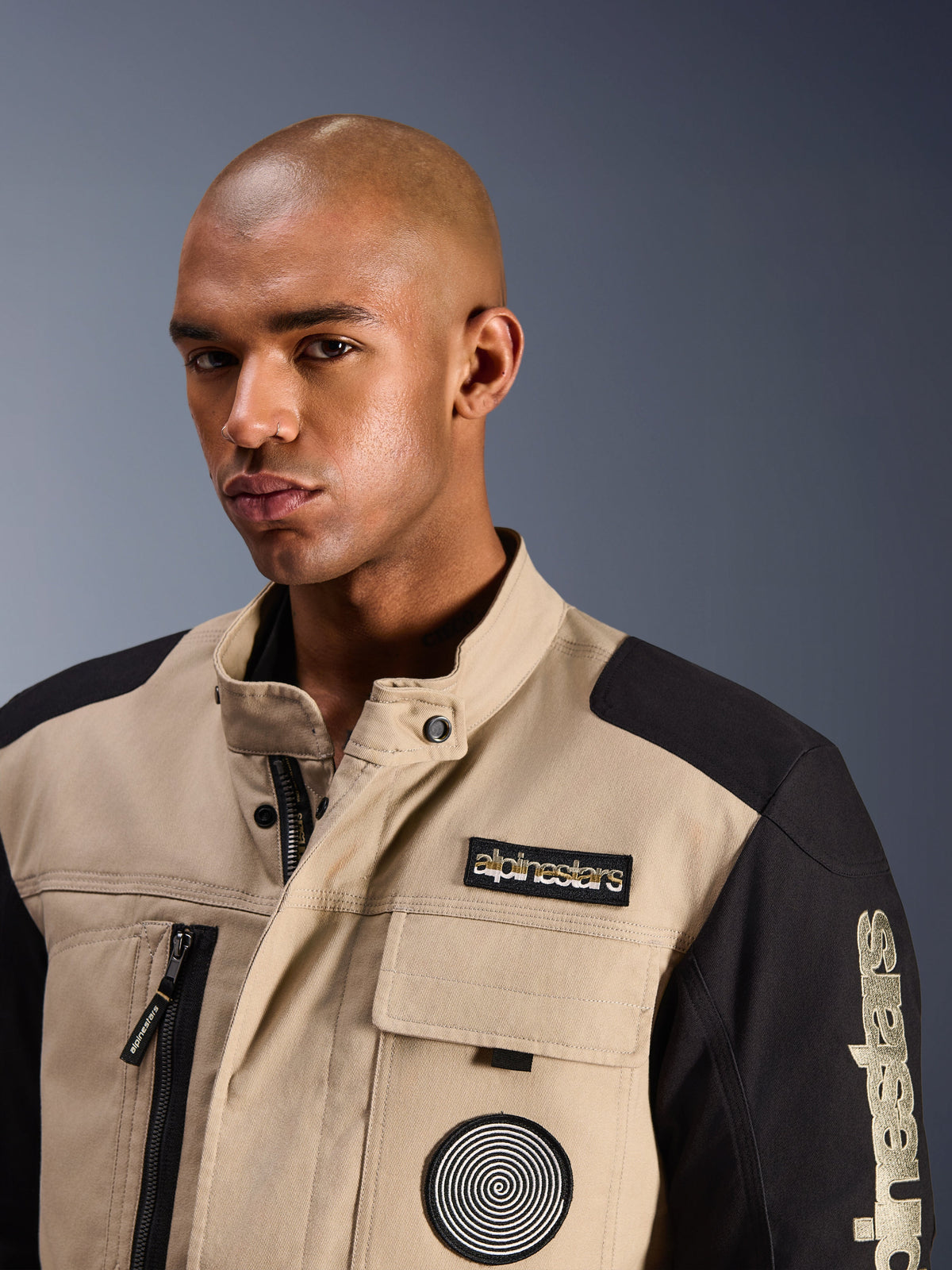 Manlig modell iförd Alpinestars Dice Jacket i Aluminum Black, en urban motorcykeljacka med färgblockerade paneler i beige och svart, med ett broderat Alpinestars-märke på bröstet och en spirallogotyp, robust arbetsklädesinspirerad design med hög krage.