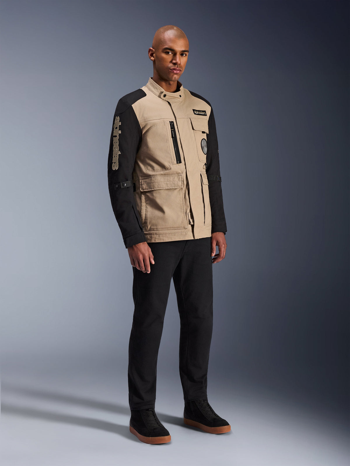 Alpinestars Dice Jacket, urban motorcykeljacka, Aluminum Black och khaki, tvåtonad design med svarta ärmar och khakifärgad torso, bärs av en modell med svarta byxor och svarta sneakers, arbetsklädesinspirerad estetik med praktiska fickor och broderad logotyp på ärmen.