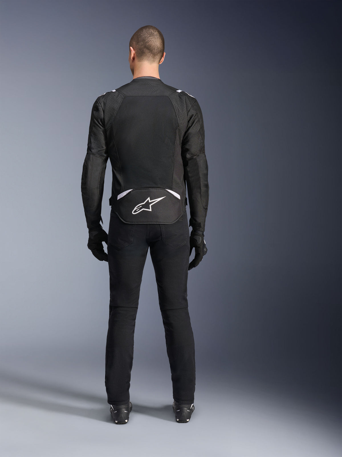 Alpinestars T-Sps Air V2-jacka, sedd bakifrån, svart och vit motorcykeljacka i textil, med en stor meshpanel på ryggen för maximalt luftflöde och vit Astars-logotyp på nedre ryggen, bärs med svarta mc-byxor och handskar