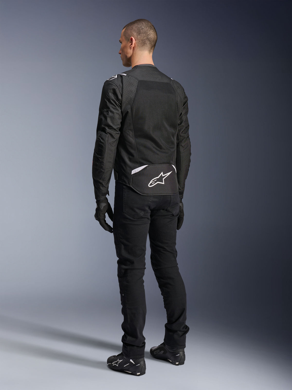 Alpinestars T-Sps Air V2-jacka, textil motorcykeljacka, svart, baksida som visar stor ventilationspanel i mesh och vit Alpinestars-logotyp på nedre delen av ryggen, kombinerad med svarta mc-byxor och handskar för en sportig urban stil.