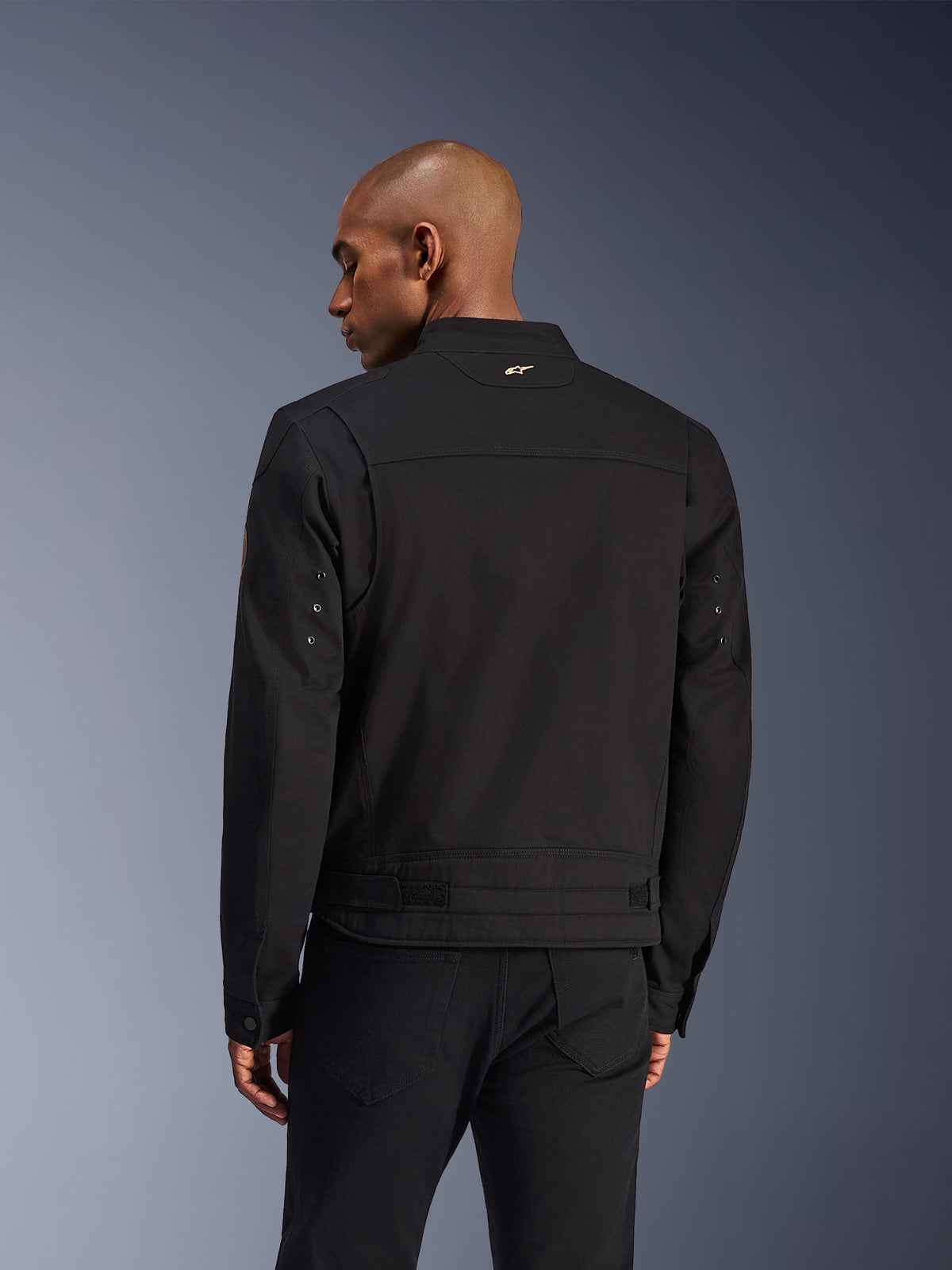 Alpinestars Bruiser-jacka, svart urban motorcykeljacka, baksida, med en arbetsklädesinspirerad design, liten guldlogotyp på kragen och justerbara midjeremmar, slitstark textilkonstruktion för stadskörning