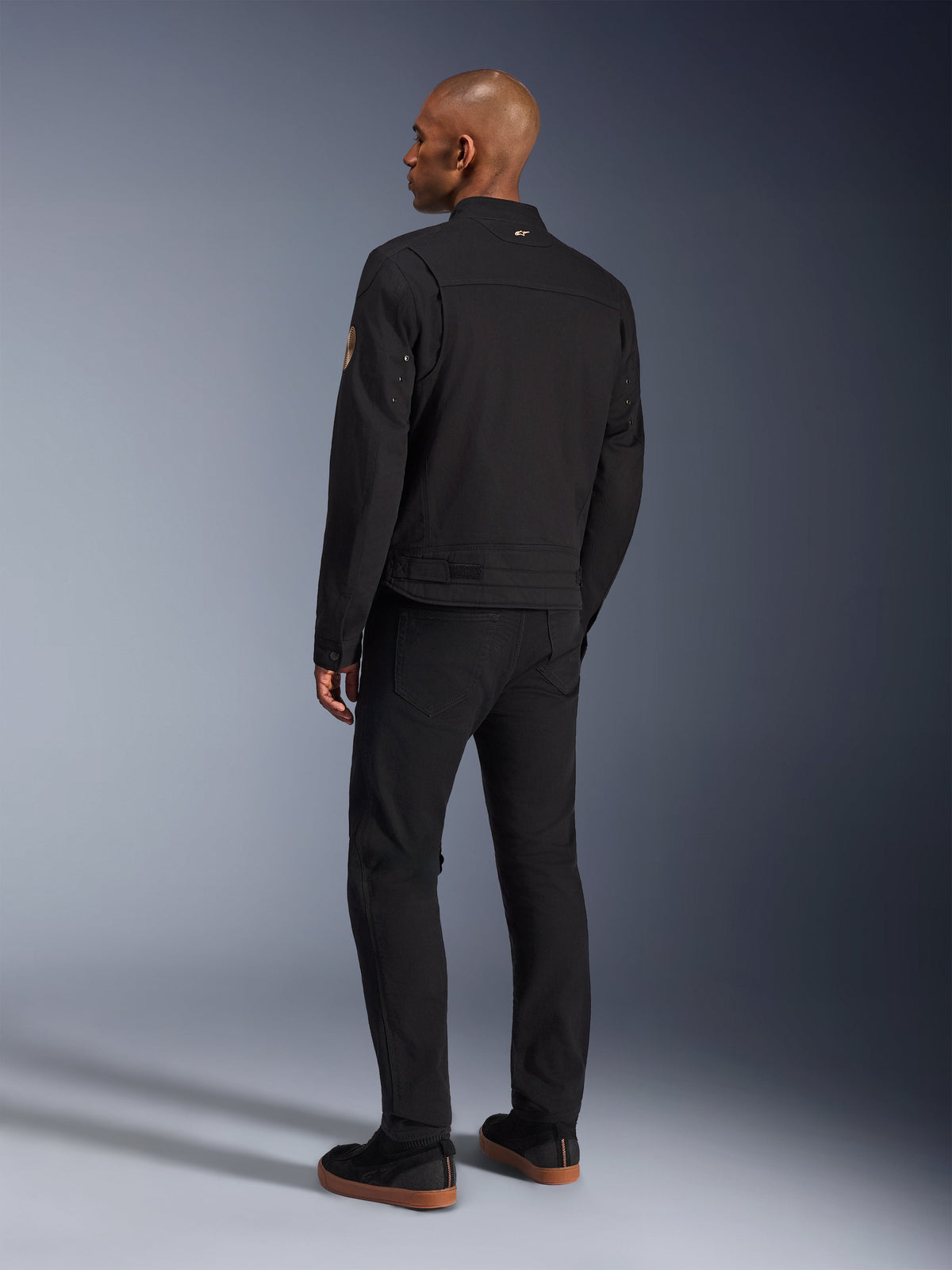 Alpinestars Bruiser-jacka, urban svart motorcykeljacka, sedd bakifrån på en modell, klassisk arbetsklädesstil, med veckade axelpaneler och Oscar-spiralmärke på ärmen, bärs med svarta byxor och avslappnade svarta skor med gummisulor.