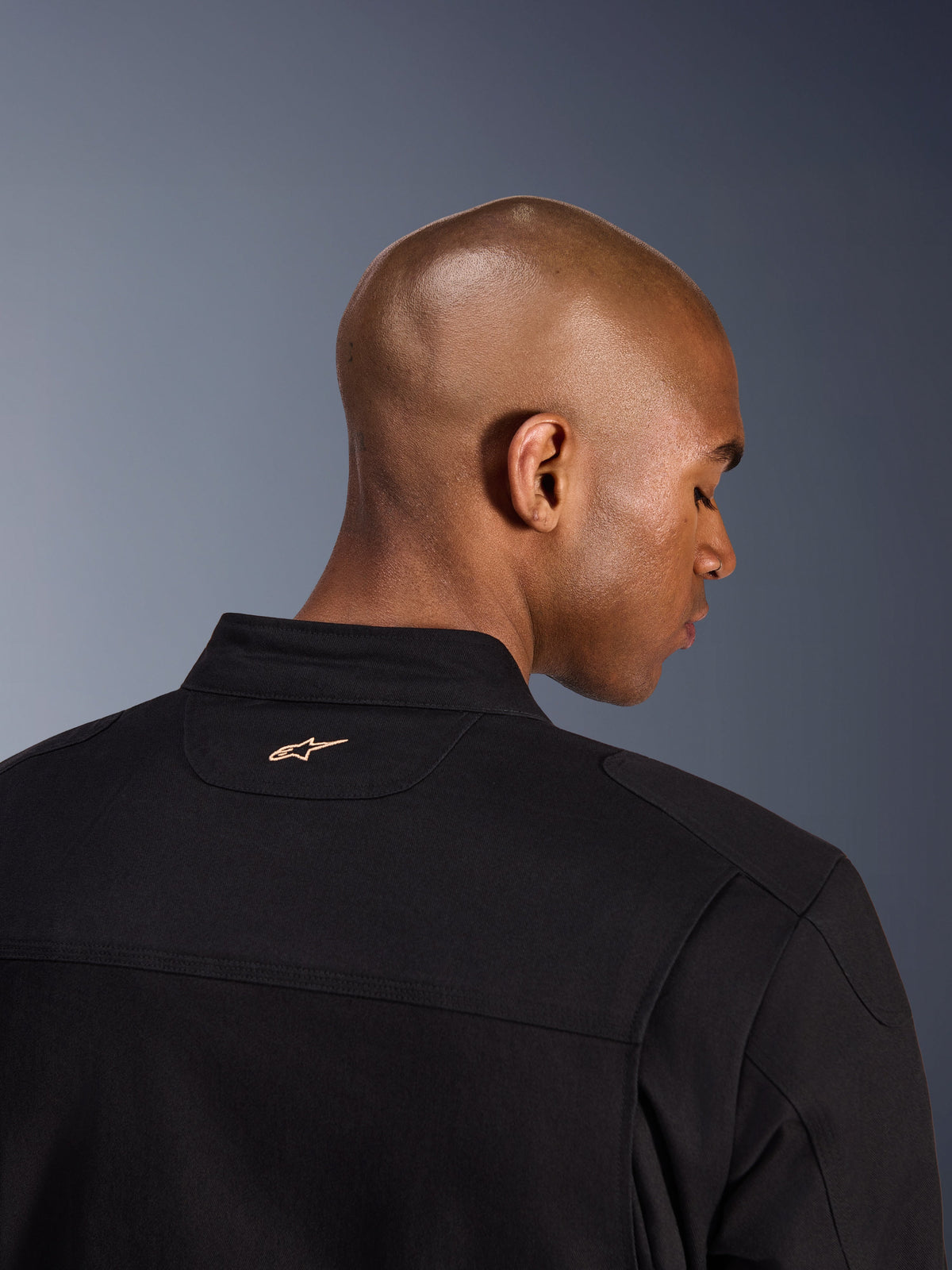 Alpinestars Bruiser Jacka, svart, detaljbild bakifrån på krage och övre axlar, liten beige broderad Alpinestars-logotyp i nacken, urban motorcykeljacka med arbetsklädesestetik