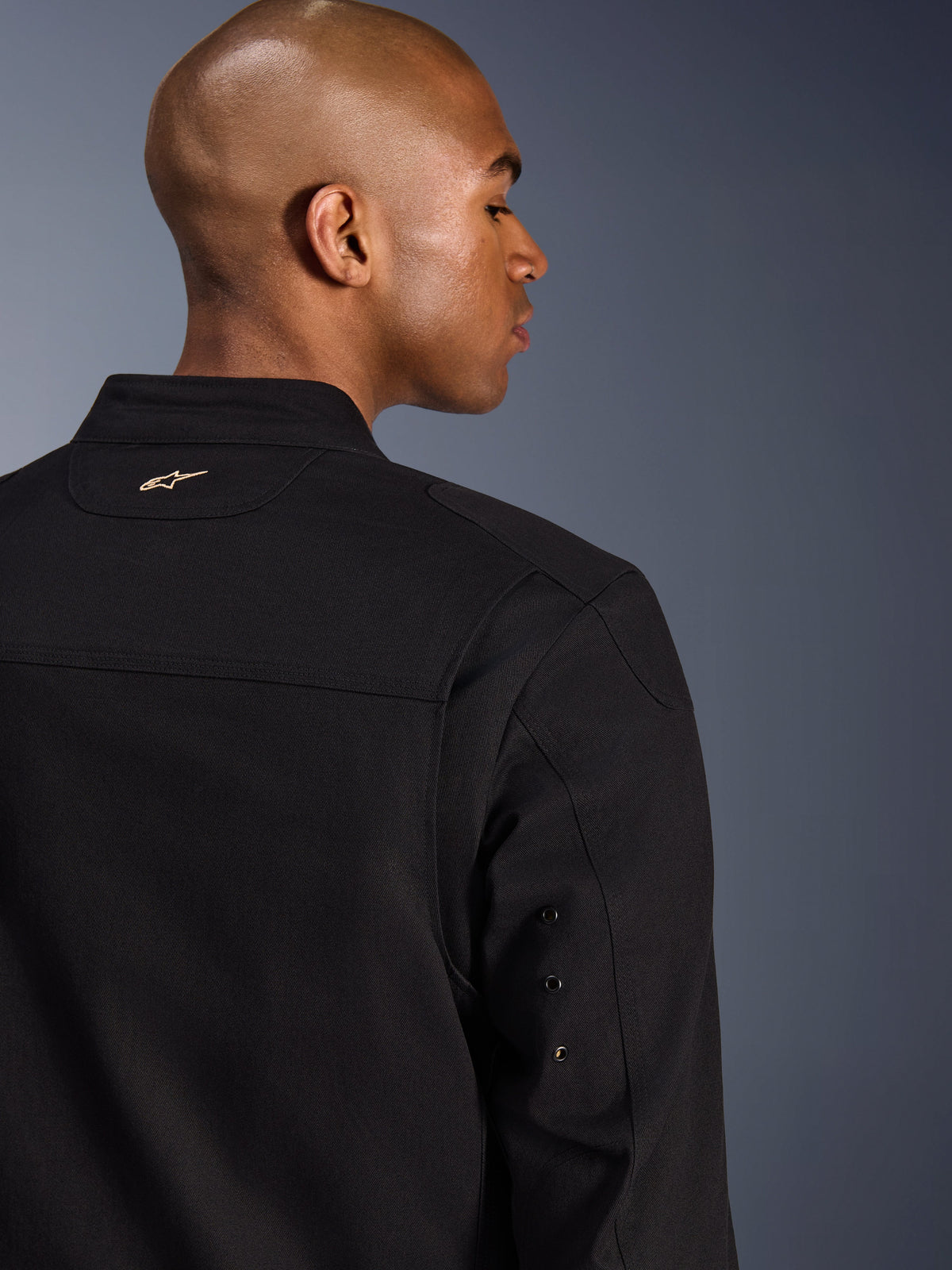 Alpinestars Bruiser Jacket, urban motorcykeljacka i textil, svart, närbild bakifrån med en broderad guldlogotyp på övre ryggen, ärmdetaljer med metallöljetter och plisserade axelpaneler för ökad rörlighet.