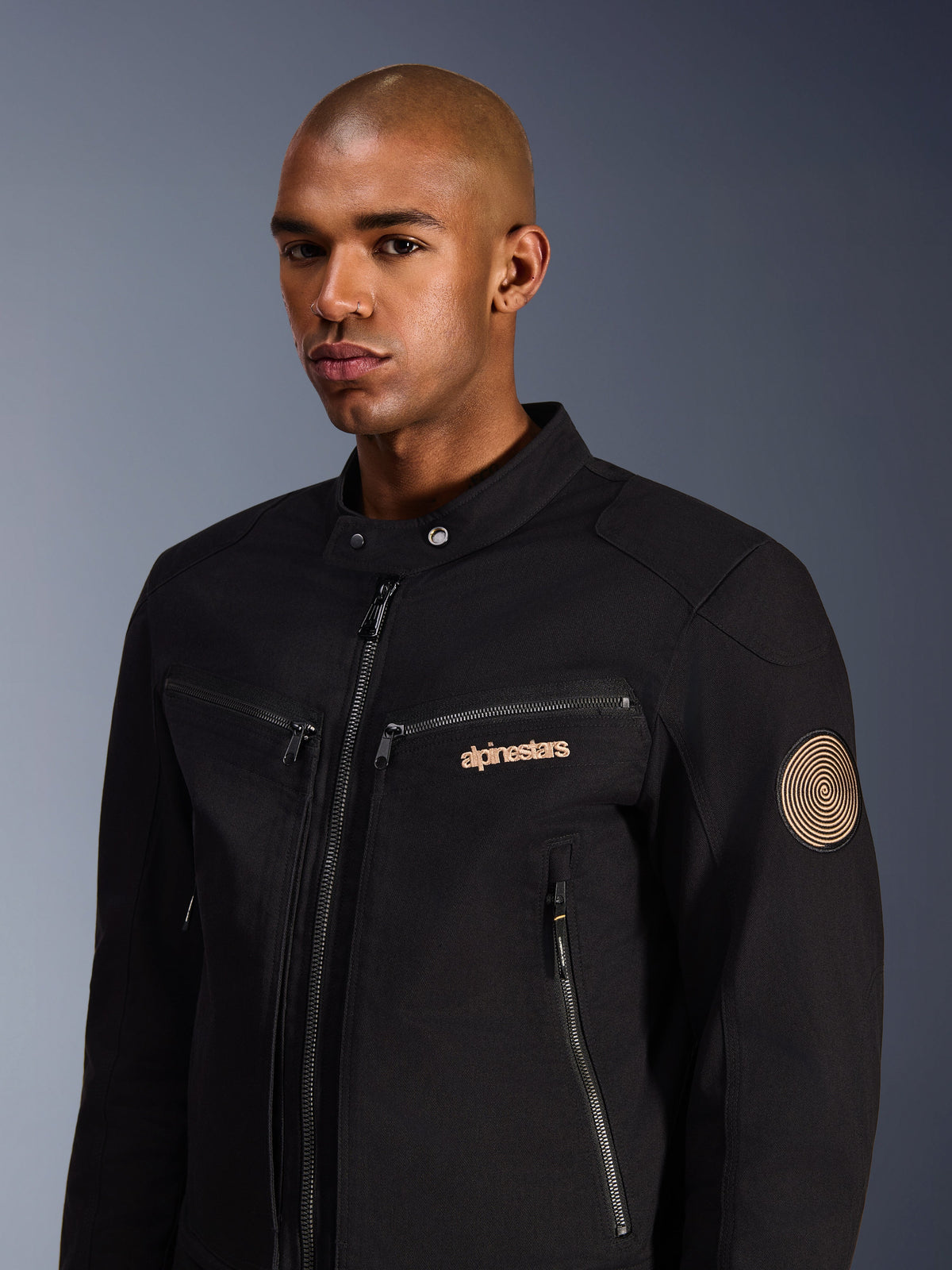 Manlig modell som bär Alpinestars Bruiser-jacka, svart, urban motorcykeljacka i polybomullstwill, med dragkedjeförsedda fickor, guldbroderad logotyp på bröstet och Oscar-spiralpatch på ärmen, modern pendlingsstil