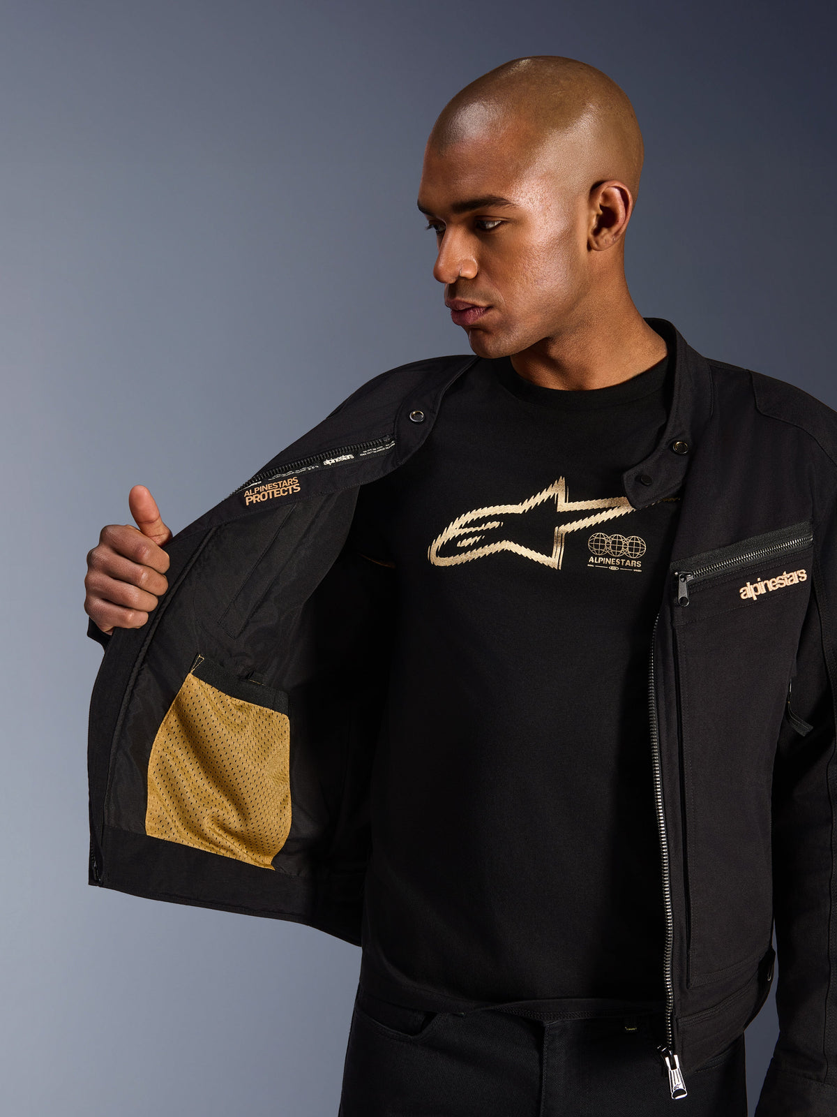 Alpinestars Bruiser-jacka, svart urban motorcykeljacka, närbild som visar den öppna insidan med guldfärgad meshficka och 'Alpinestars Protects'-logotyp, buren över en svart t-shirt med logotyp, textil mc-utrustning med arbetsklädesestetik