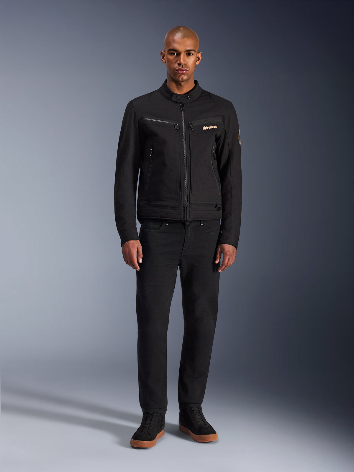 Alpinestars Bruiser Jacket, svart urban motorcykeljacka, bärs av modell med svarta byxor och high-top sneakers, vy framifrån som visar bröstfickor med dragkedja och broderad logotyp, klassisk arbetsklädesinspirerad design för daglig pendling