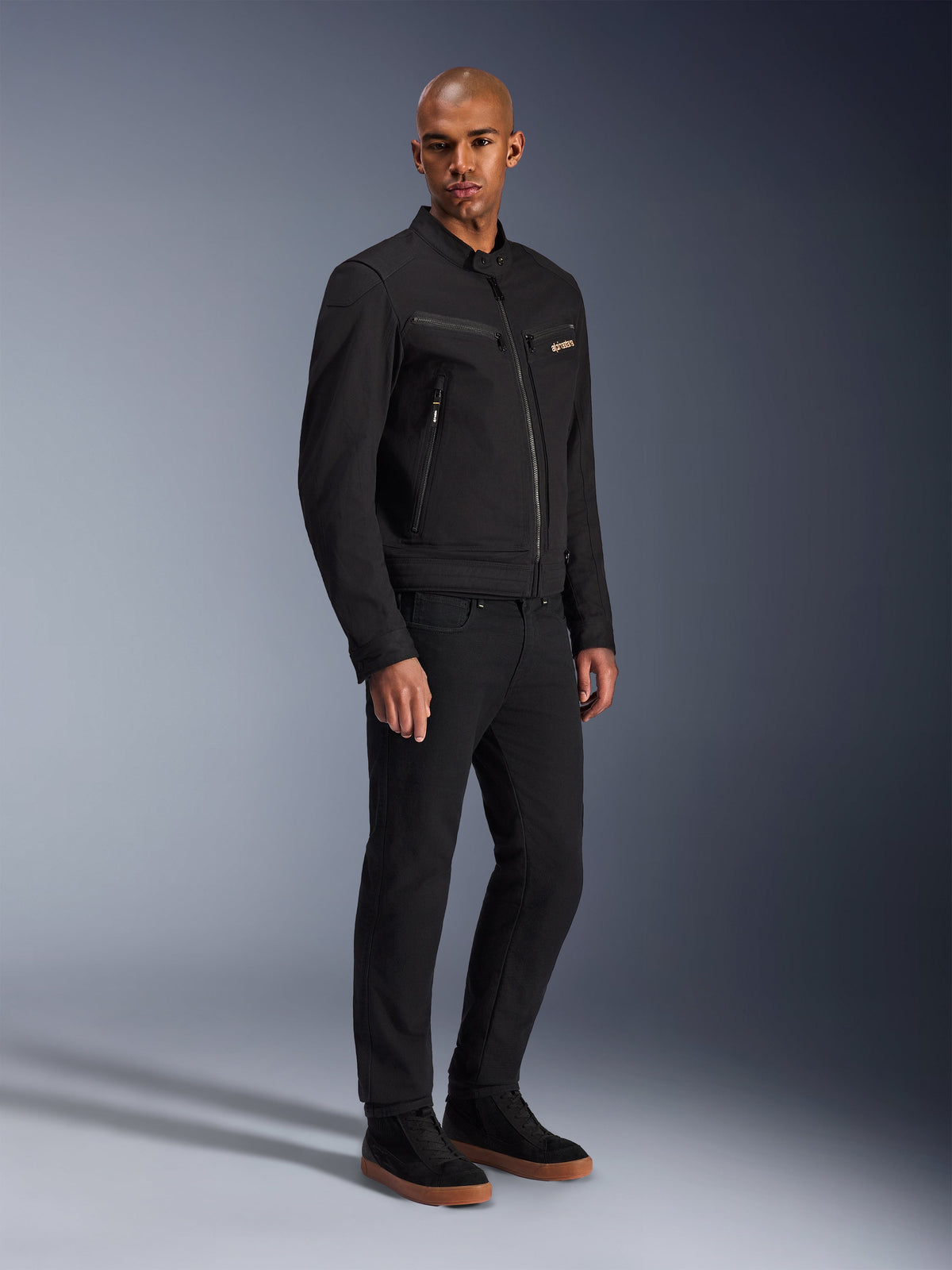 Alpinestars Bruiser Jacket, urban motorcykeljacka, svart, bärs av en modell med svarta byxor och sneakers, med bröstfickor med dragkedja, polybomullstwill och en stilren arbetsklädesinspirerad design för urban körning