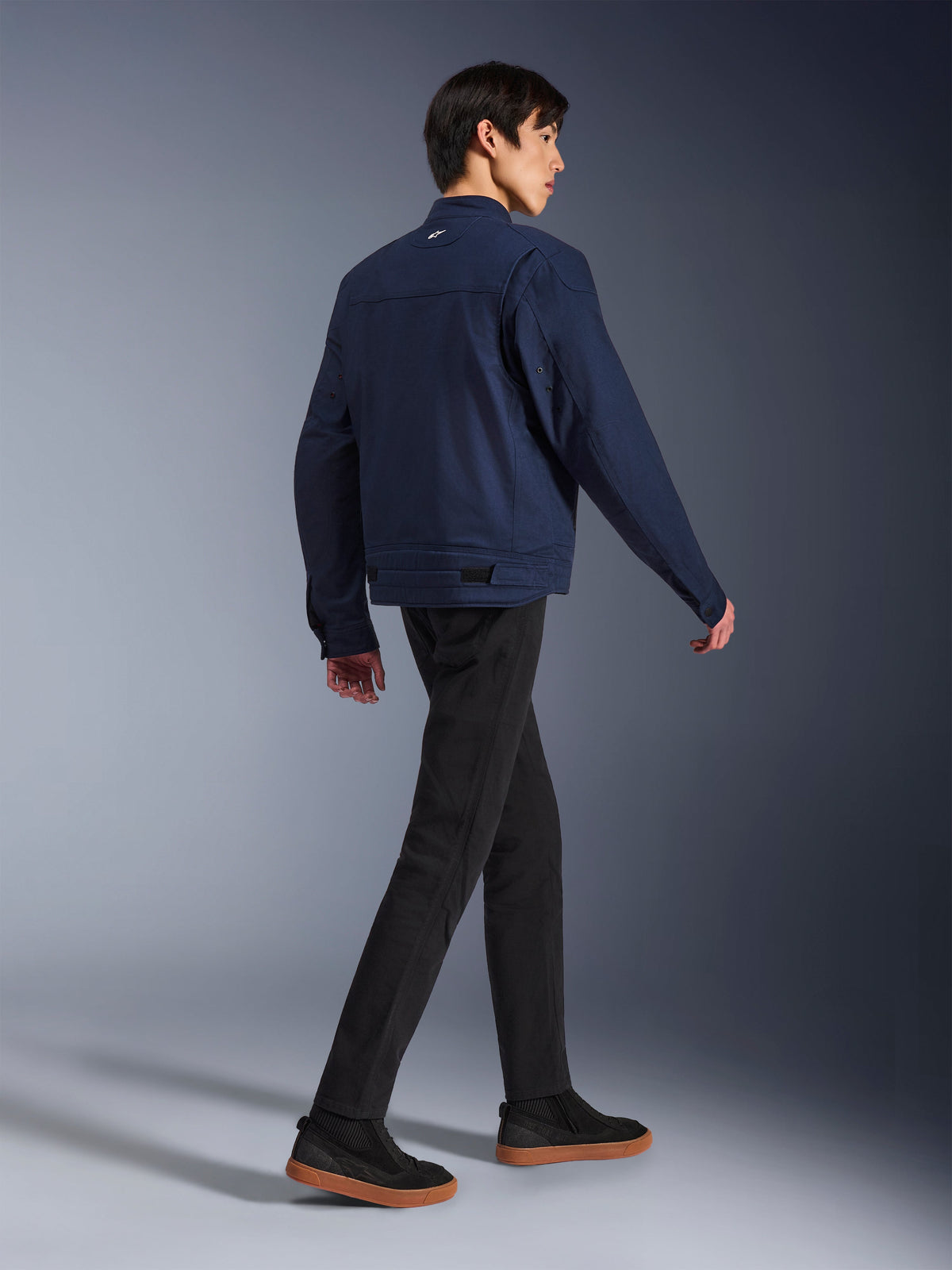 Alpinestars Bruiser-jacka i Dark Blue, urban motorcykeljacka i textil, sedd bakifrån på gående modell, kombinerad med svarta byxor och sneakers med gummisula, med arbetsklädesinspirerad design och justerbara flikar i midjan