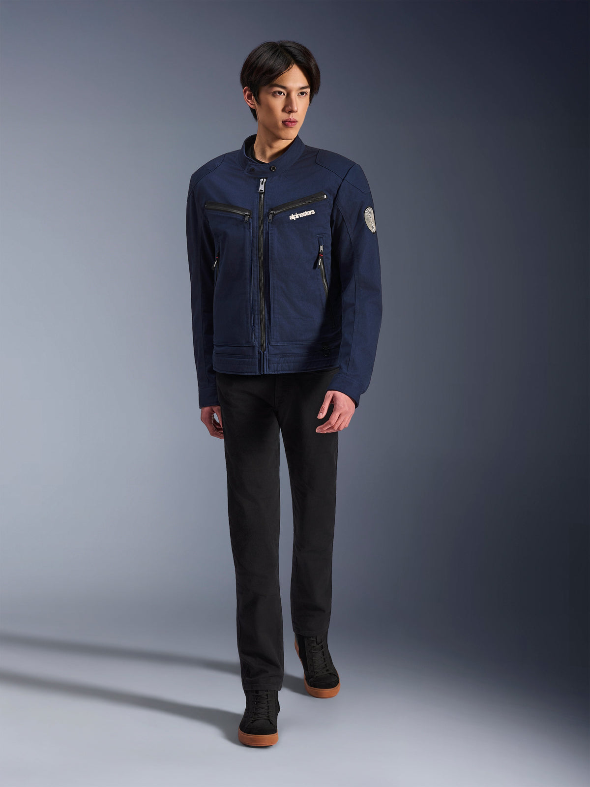 Manlig modell bär Alpinestars Bruiser Jacket i mörkblått, urban mc-jacka i textil, matchad med svarta byxor och svarta sneakers, arbetsklädesinspirerad estetik med dragkedjefickor och ett cirkulärt märke på ärmen, modern streetwear för motorcykelkörning