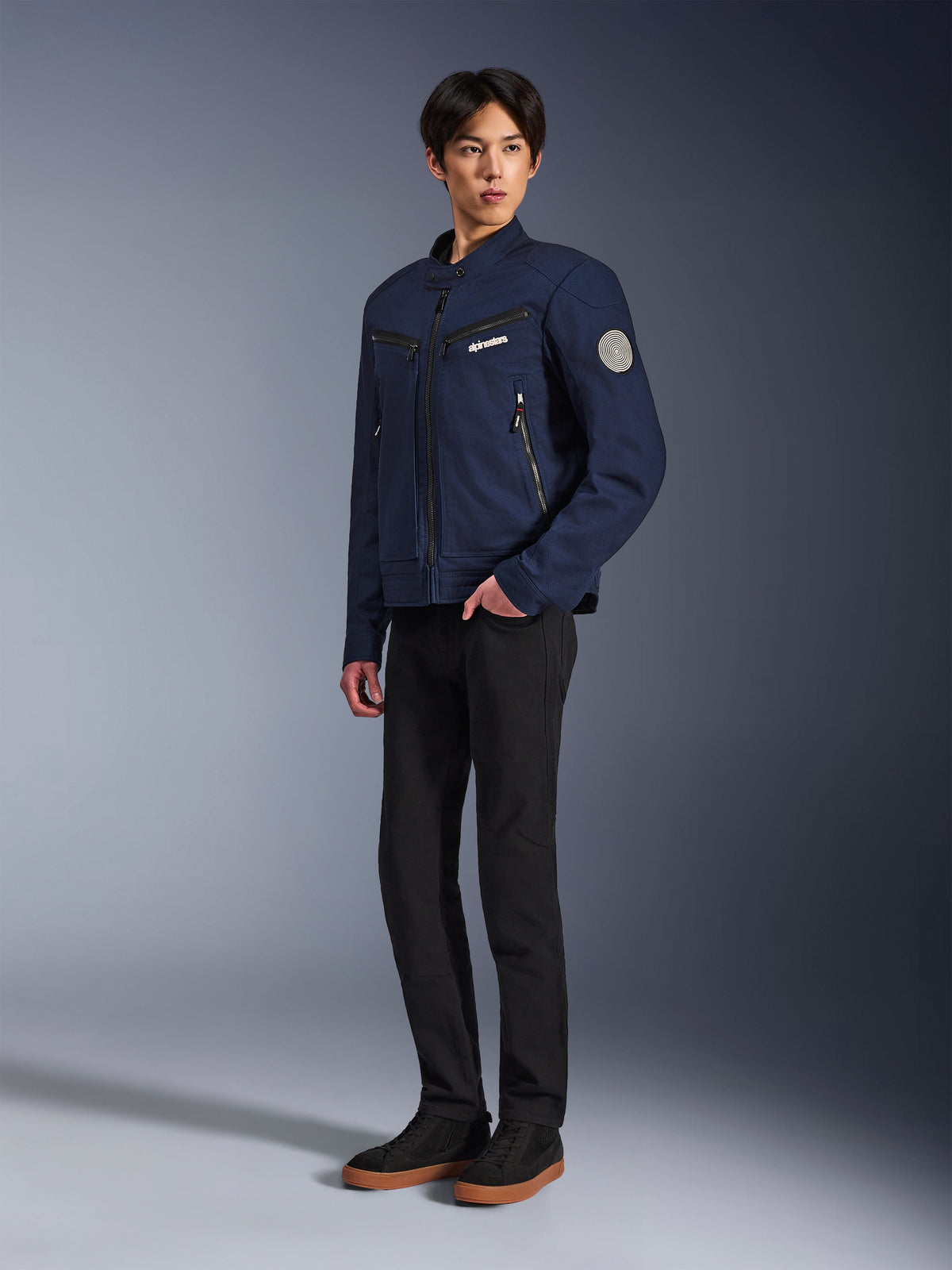 Alpinestars Bruiser Jacka i mörkblått, urban motorcykeljacka i textil med arbetsklädesinspirerad design, med bröstfickor med dragkedja och Oscar-spiralmärke på ärmen, bärs med svarta byxor och avslappnade sneakers.