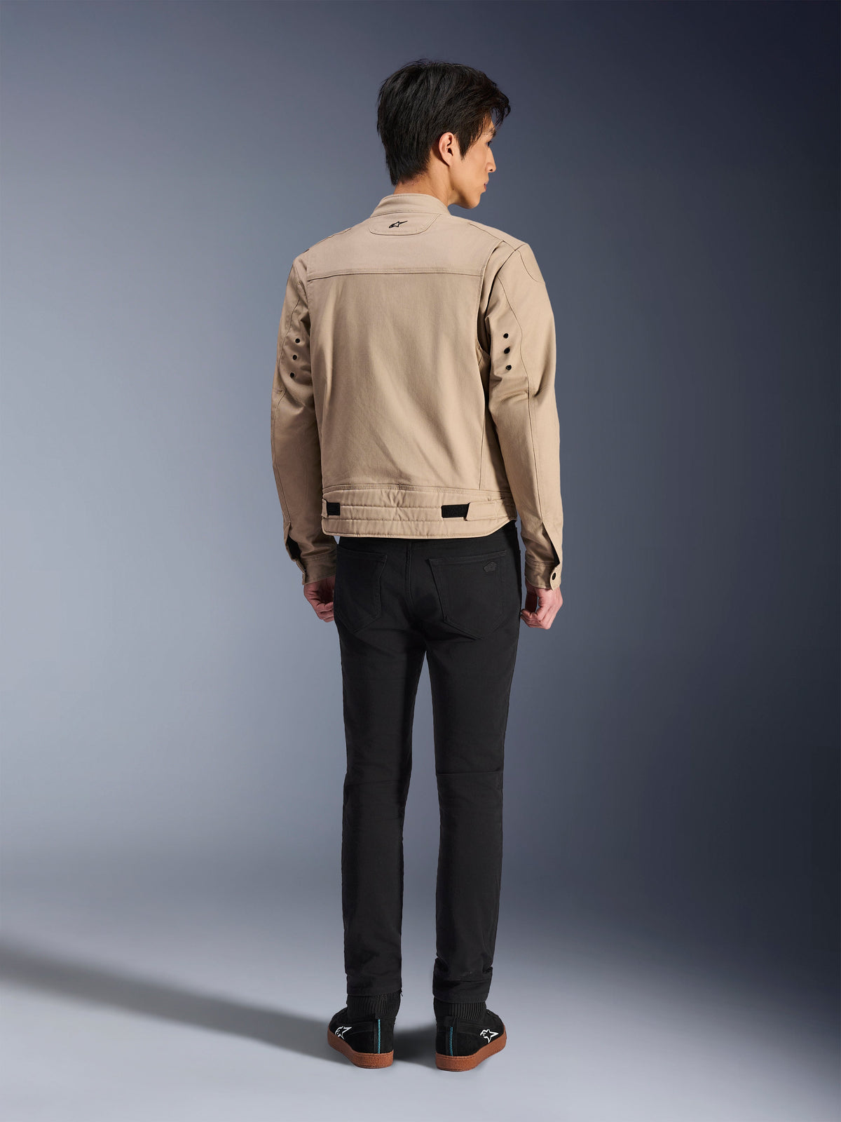 Ryggvy av en modell som bär Alpinestars Bruiser Jacket i brunt, en urban motorcykeljacka med arbetsklädesinspirerad design, med detaljer av metallöljetter på ryggen och midjejusteringar, kombinerad med svarta byxor och sneakers, modern urban körstil.
