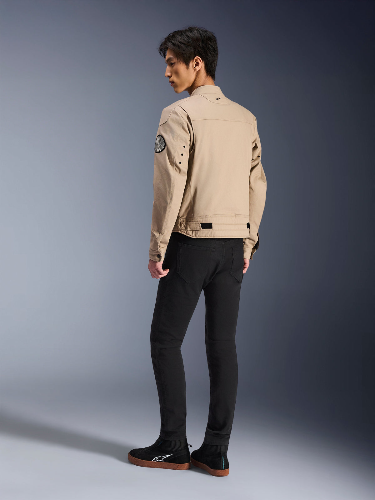 Manlig modell sedd bakifrån bärande Alpinestars Bruiser Jacket i tan, urban modern motorcykeljacka med arbetsklädesstil, med ett cirkulärt märke på ärmen, midjejusteringsremmar och artikulerade axlar, kombinerat med svarta byxor och svarta sneakers.
