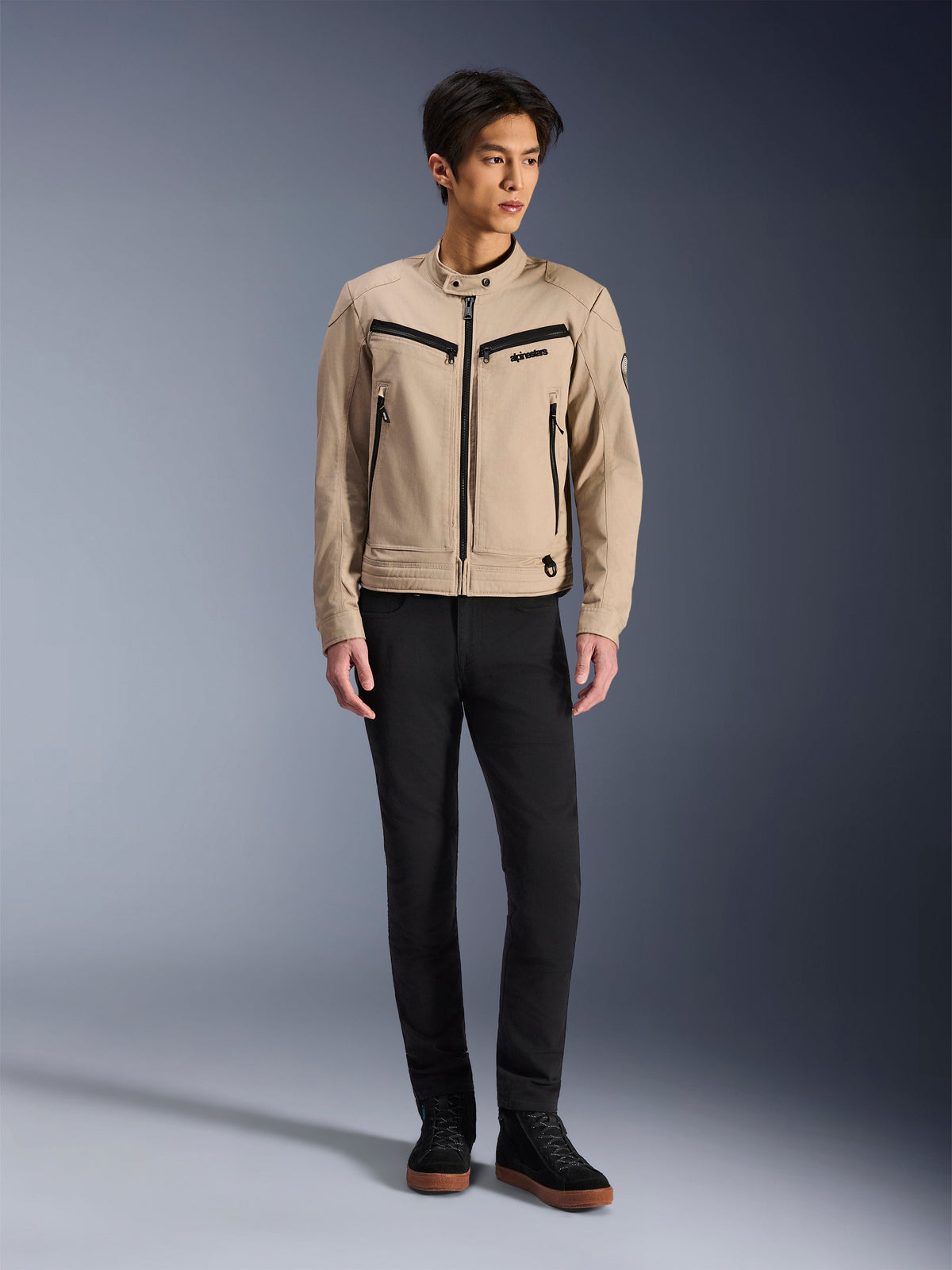 Alpinestars Bruiser Jacket, urban motorcykeljacka i Aluminum, bärs med svarta byxor och svarta sneakers, med svarta dragkedjefickor och krage med tryckknapp, modern arbetsklädesinspirerad design för stadskörning