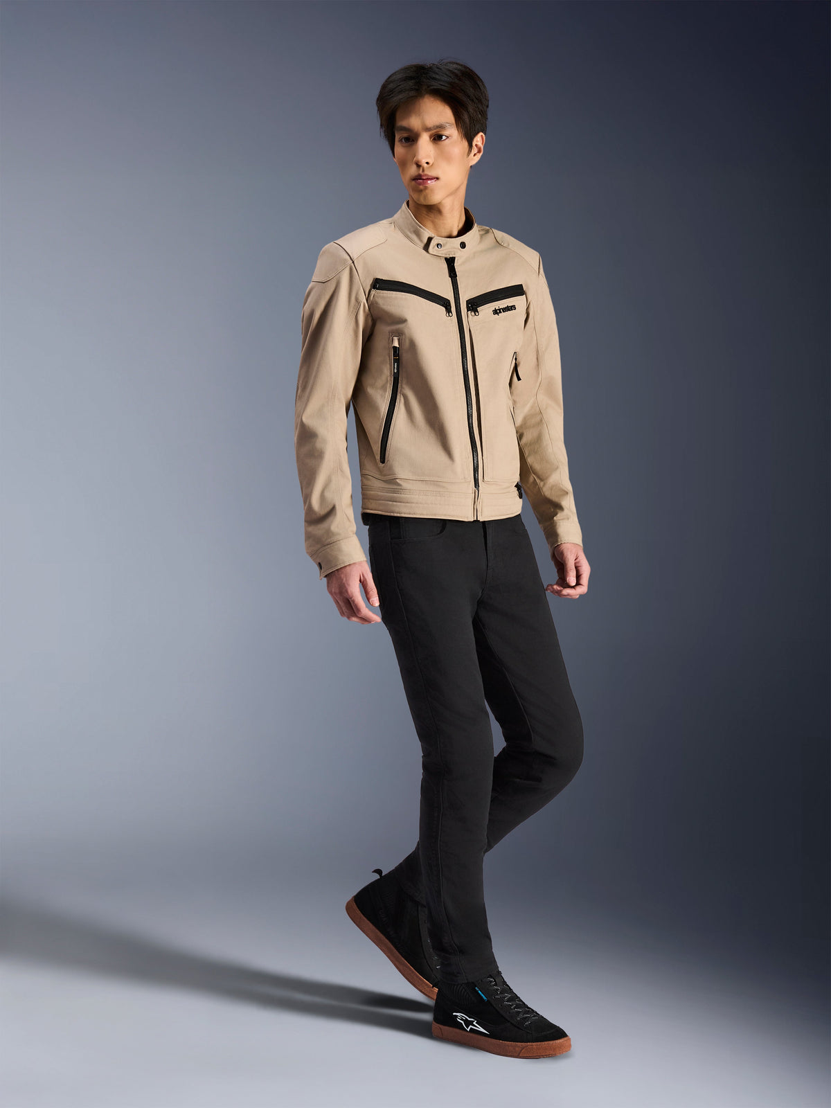 Alpinestars Bruiser-jacka, urban motorcykeljacka, tan, bärs av en manlig modell med svarta slim-fit-byxor och svarta sneakers, arbetsklädesinspirerad textilkonstruktion med svarta dragkedjedetaljer, samtida urban street-stil.