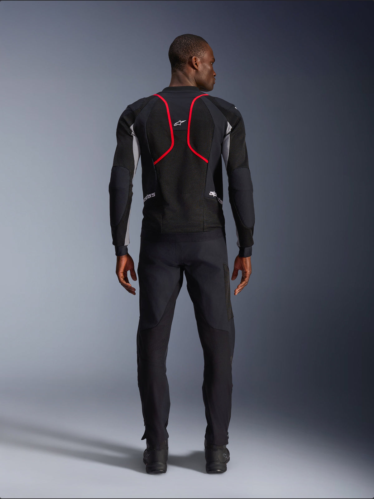Alpinestars Troop-Air Pro Jacket, svart med detaljer i Ash Gray och Bright Red, modell sedd bakifrån, teknisk mesh-konstruktion med framträdande röd piping på ryggen, Alpinestars-logotyp upptill på ryggen, kombinerad med svarta motorcykelbyxor, premium bepansrad air-jacka för landsvägskörning