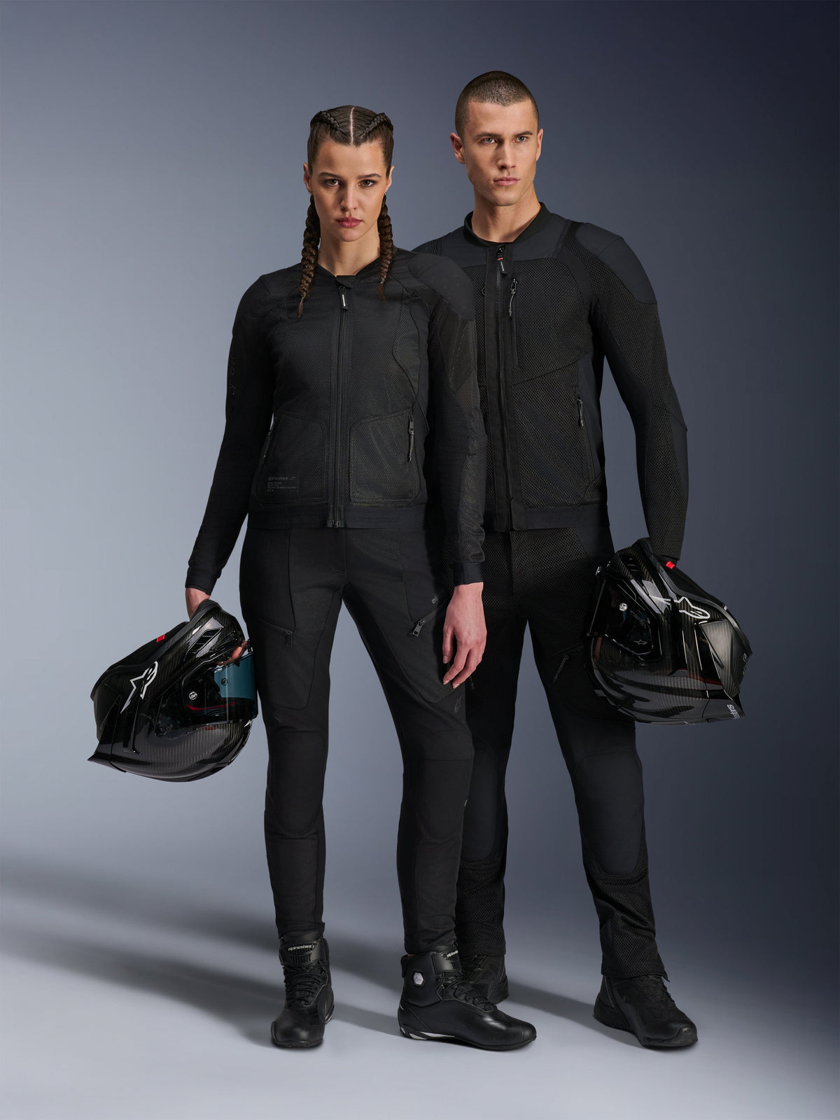 Manlig och kvinnlig modell bär svarta Alpinestars Troop-Air Pro-jackor, teknisk skyddsutrustning i mesh kombinerat med matchande svarta byxor och stövlar, båda hållande en svart hjälm, vilket visar upp koordinerade urbana motorcykelkläder.
