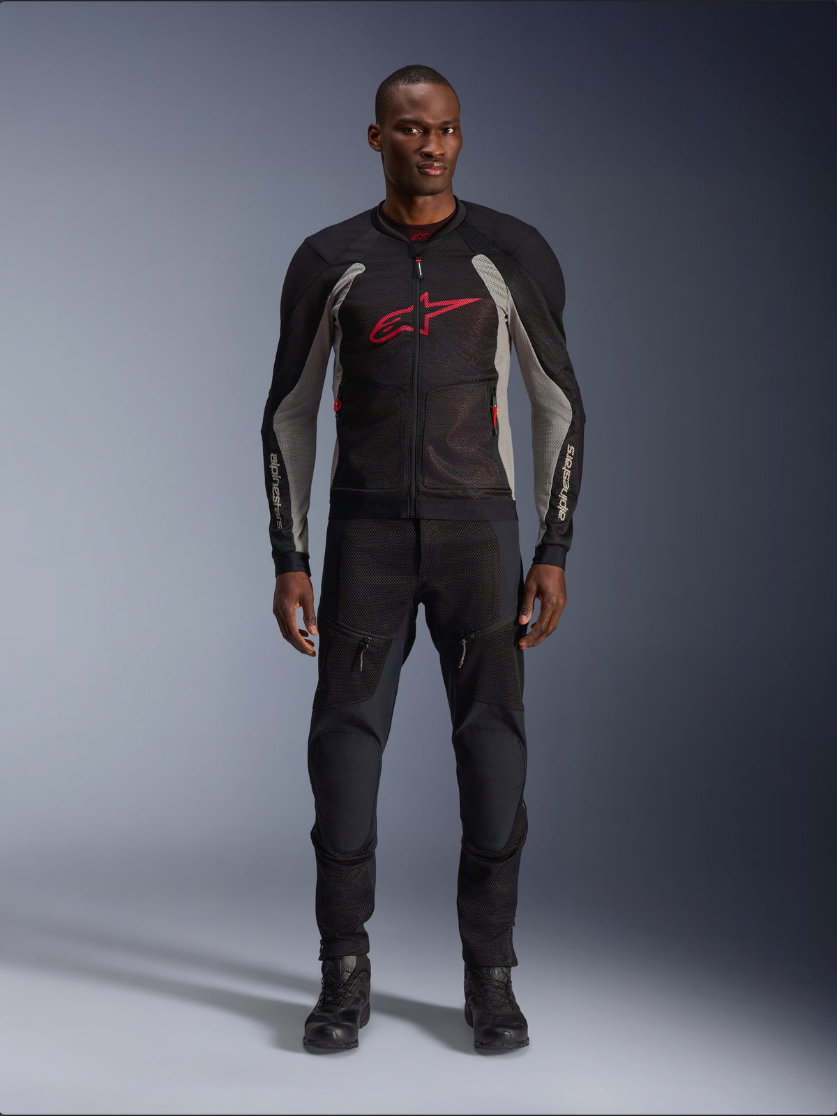 Manlig modell som bär Alpinestars Troop-Air Jacket i färgerna Black Ash Gray och Bright Red, teknisk motorcykeljacka i mesh med en framträdande röd logotyp på bröstet, stylad med svarta motorcykelbyxor och stövlar, urban mc-utrustning