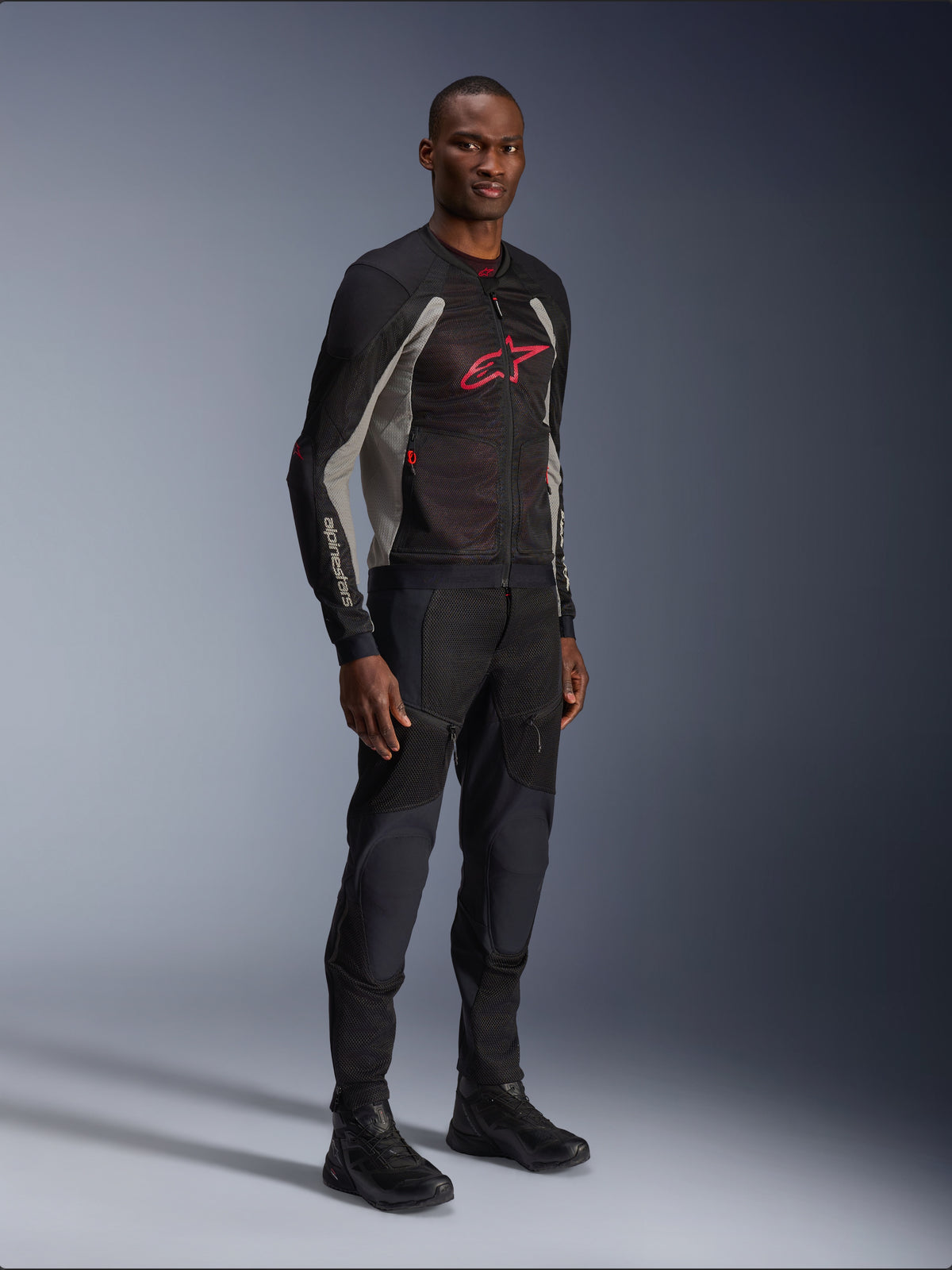 Alpinestars Troop-Air-jacka, teknisk motorcykeljacka i mesh i svart, askgrått och klar rött, bärs av en modell med matchande svarta byxor och stövlar, med en röd Alpinestars-logotyp på bröstet, urban modern mc-utrustning.