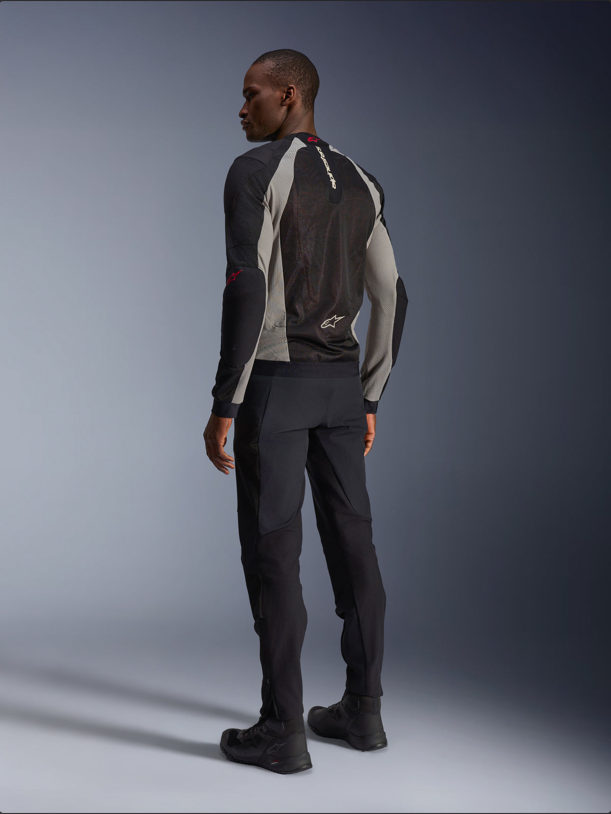 Modell klädd i Alpinestars Troop-Air-jacka sedd bakifrån, Black Ash Gray med Bright Red-detaljer, teknisk mesh-konstruktion för ventilation, matchad med svarta motorcykelbyxor och körstövlar, urban modern stil