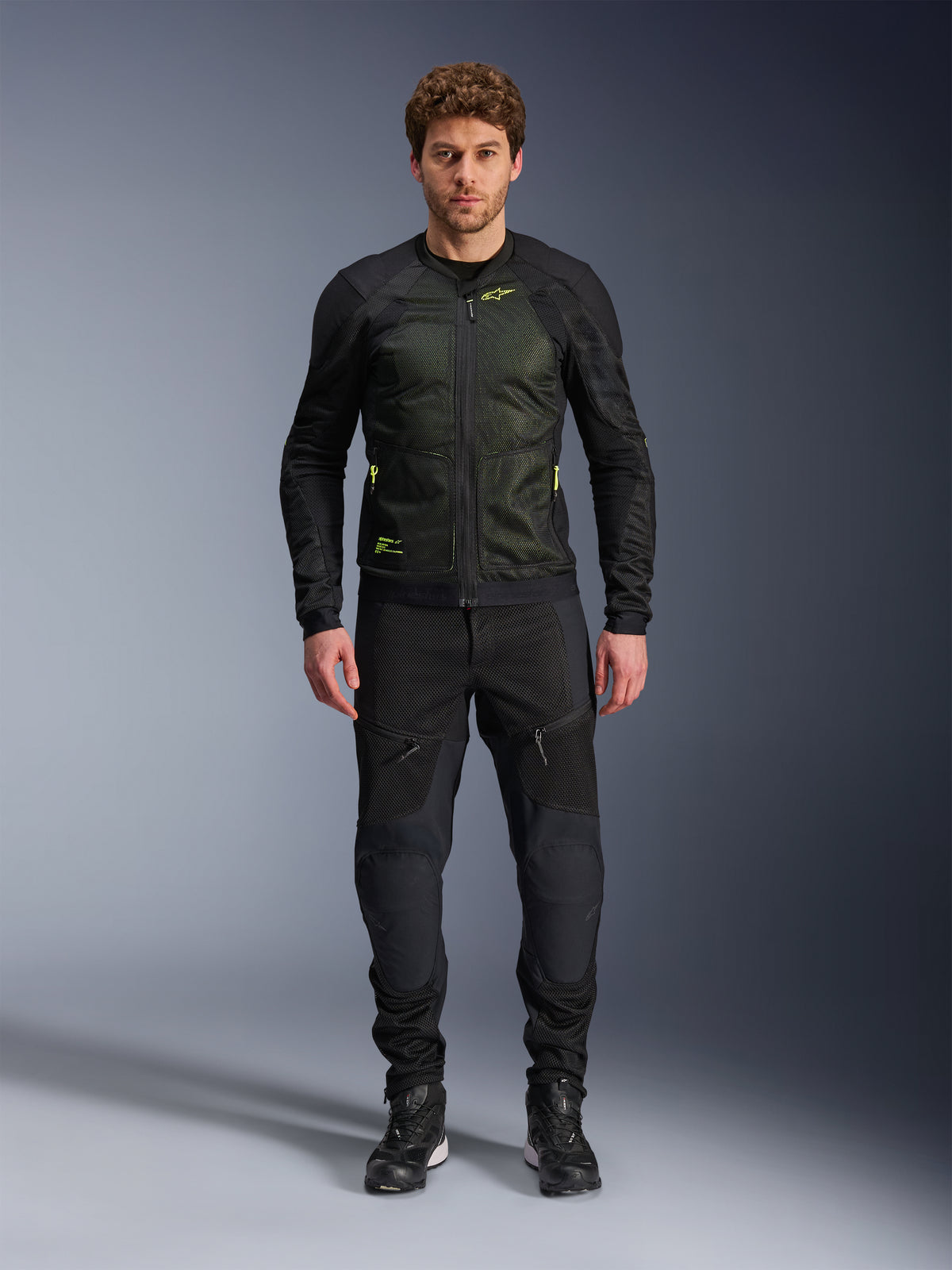 Manlig modell som bär Alpinestars Troop-Air-jacka i Black Yellow Fluo, med tekniska meshpaneler och neongula detaljer, kombinerat med svarta mc-byxor och skor, modern motorcykelutrustning för varmt väder