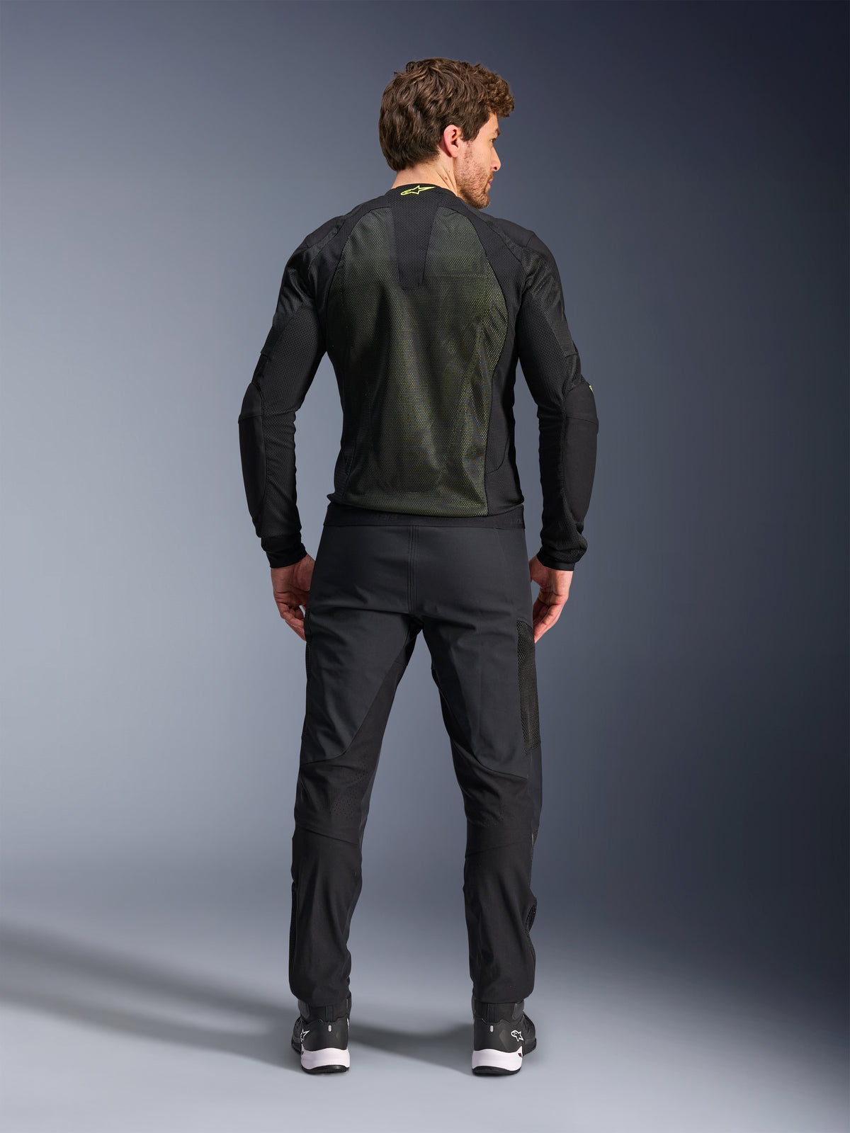 Alpinestars Troop-Air Jacka, Black Yellow Fluo, sedd bakifrån på en modell, med en stor 2D tech mesh-ryggpanel för ventilation, teknisk motorcykeljacka kombinerad med svarta körbyxor och stövlar, urban motorcykelstil