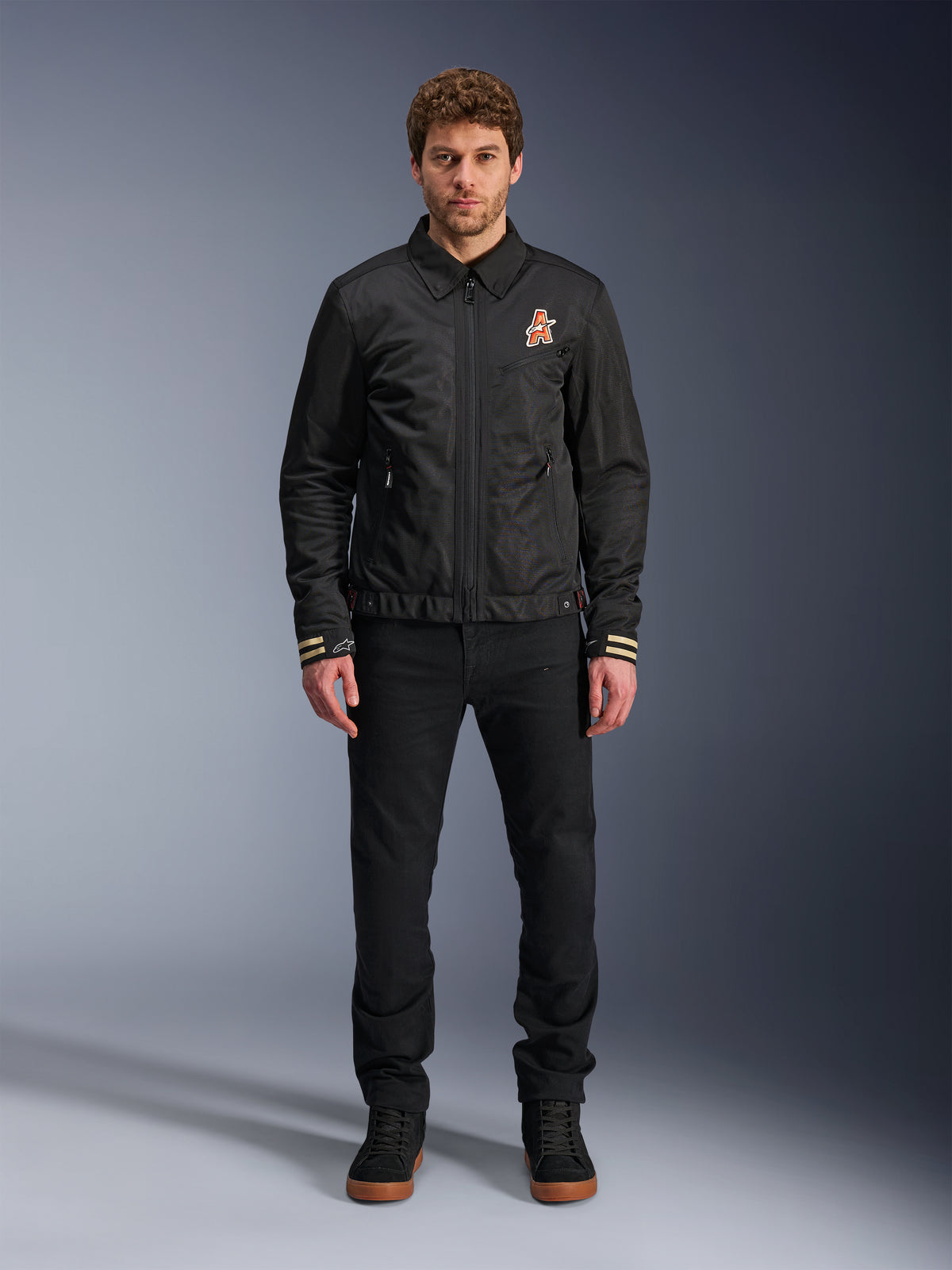 Manlig modell som bär Alpinestars Ignite Air Jacka i svart, med en klassisk skjortkrage, hel dragkedja fram och en orange logotyp på bröstet. Stylad med svarta byxor och svarta high-top sneakers för en urban, samtida motorcykelstil.