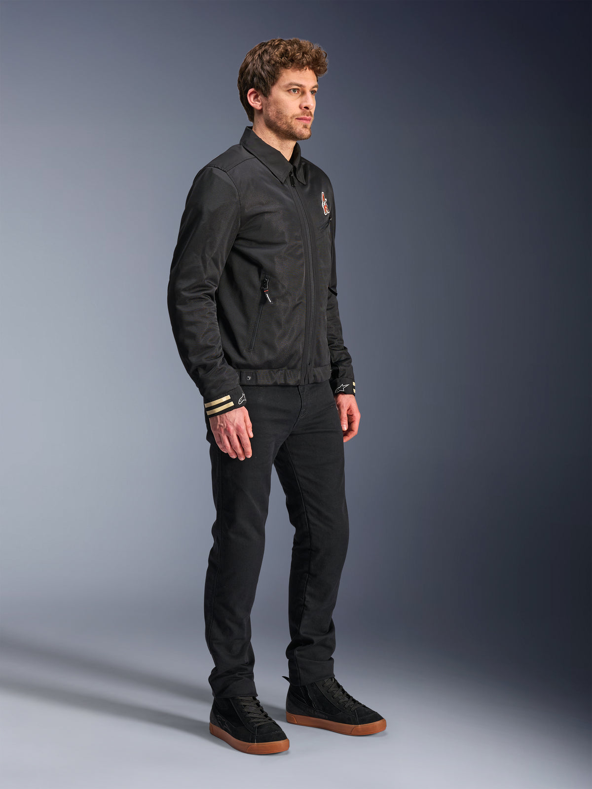 Alpinestars Ignite Air Jacket, urban motorcykeljacka, svart, bärs av en modell med svarta byxor och svarta sneakers, med traditionell skjortkrage, klubbinspirerad grafik på bröstet och ärmsluten, elegant modern design med mesh och satinfinish