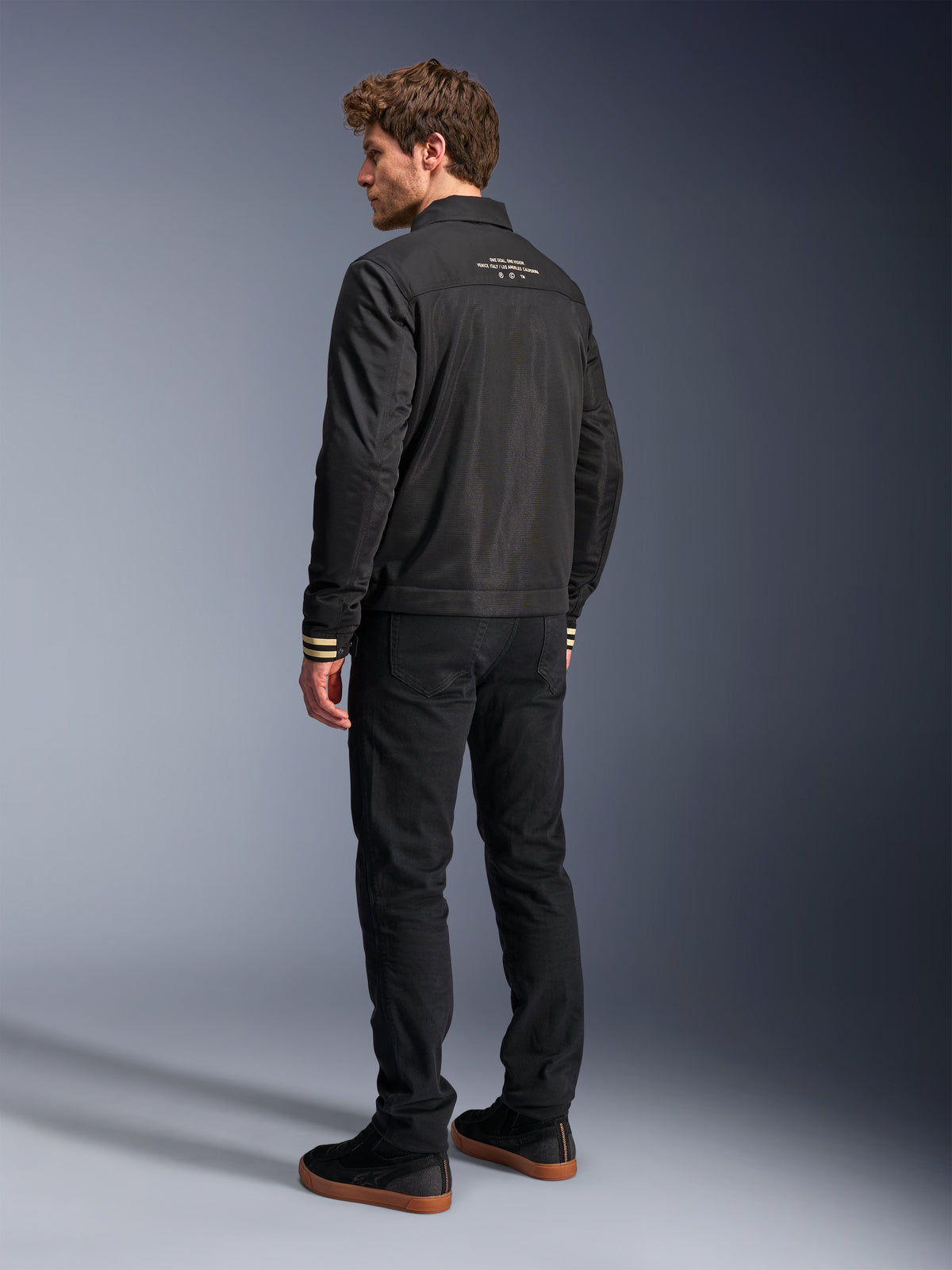 Alpinestars Ignite Air Jacka, svart motorcykeljacka, vy bakifrån med vit grafisk text på övre ryggen och beige-randiga ärmslut, Harrington-silhuett med mesh-konstruktion, matchad med svarta byxor och svarta sneakers