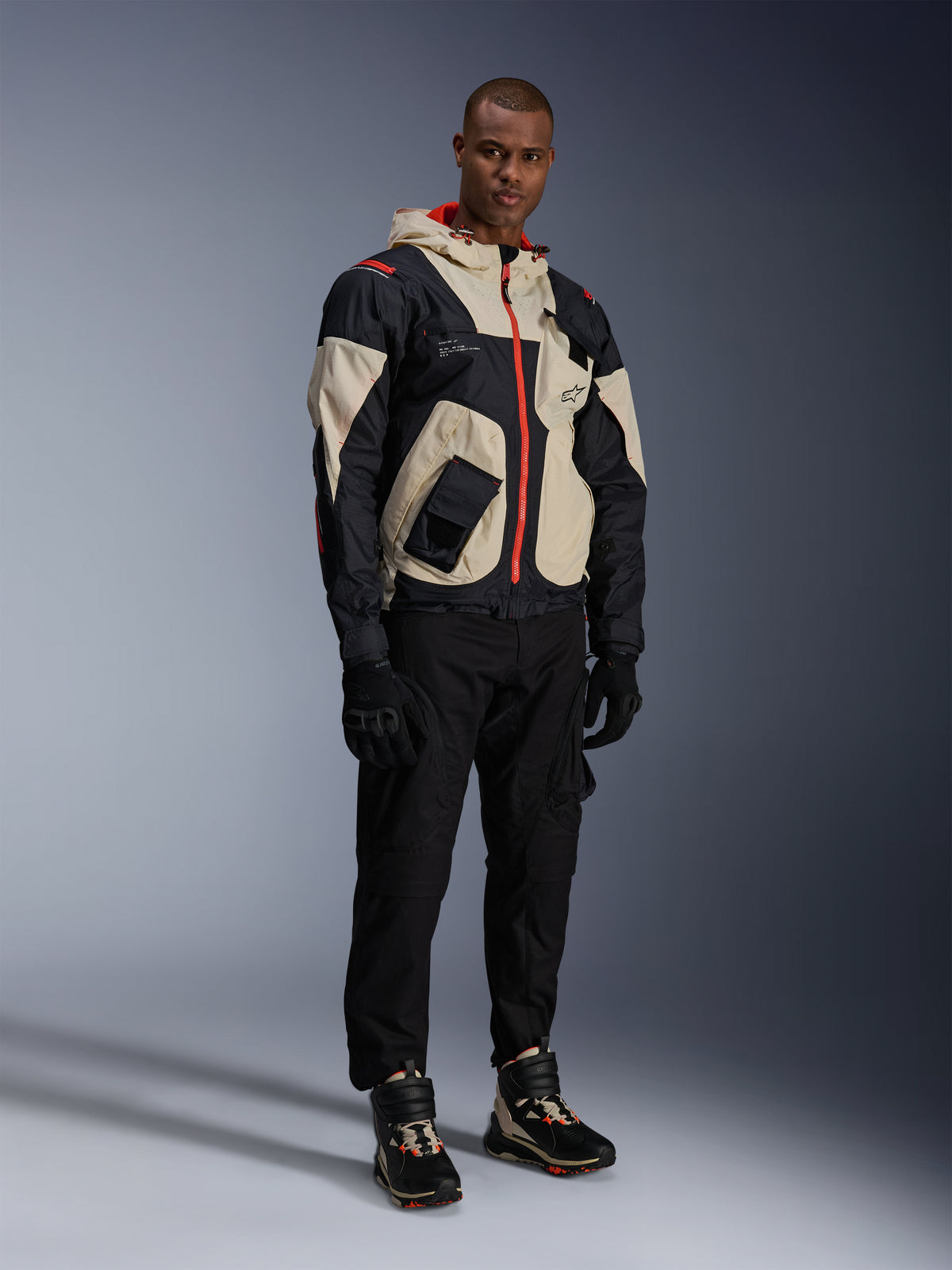 Man som står i komplett urban motorcykelutrustning, med Alpinestars Speedstreet Shoes i Black Almond Hot Orange och en Mogress Air Jacket i svart och krämfärgat, matchande svarta byxor och handskar, streetwear-inspirerad skyddsutrustning för stadskörning