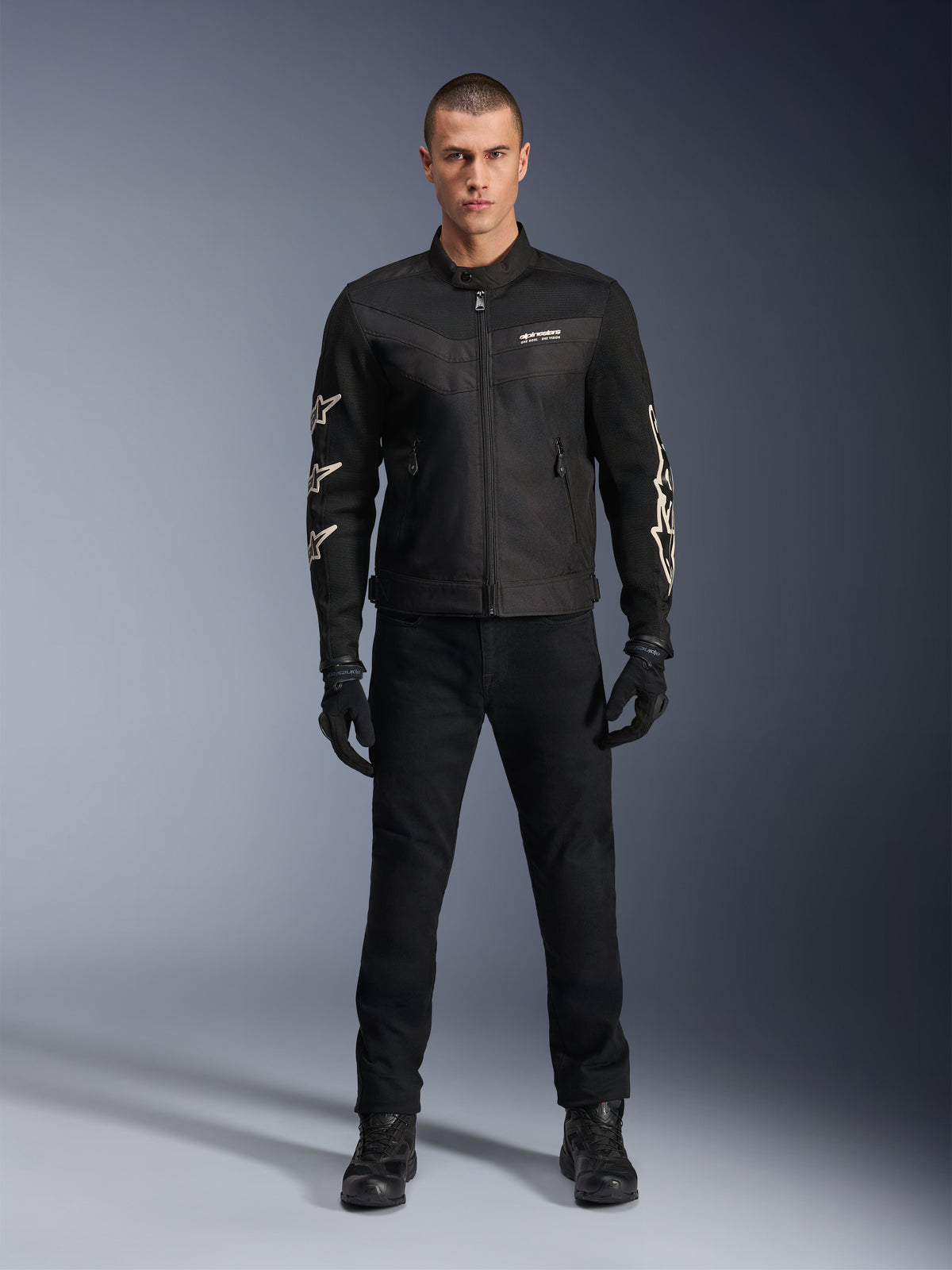 Male model wearing Alpinestars T-Dyno Air Jacket, Black, lightweight textile motorcycle jacket with breathable 3D mesh, white star logos on sleeves, paired with black pants, gloves, and boots, front view standing