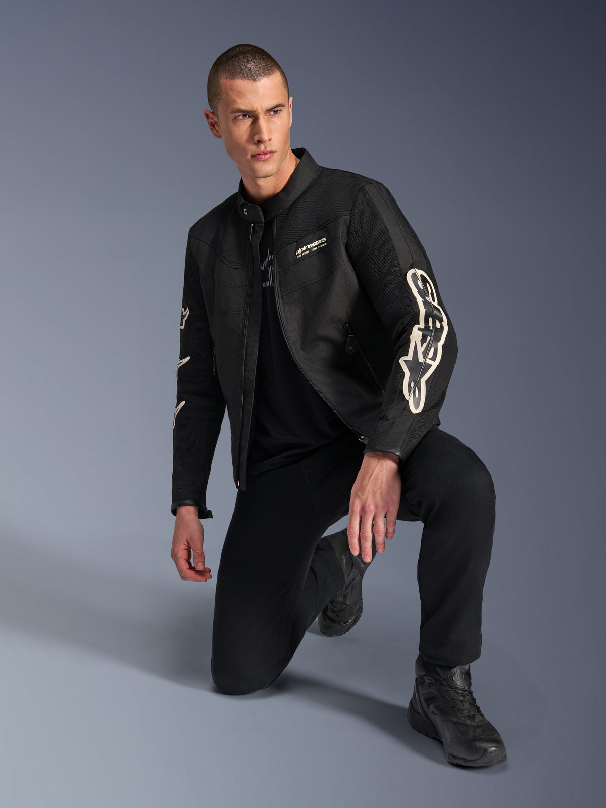 Male model wearing Alpinestars T-Dyno Air Jacket, Black textile motorcycle jacket with large white branding on sleeves, paired with black pants and riding boots, modern vintage-inspired design with 3D mesh panels.