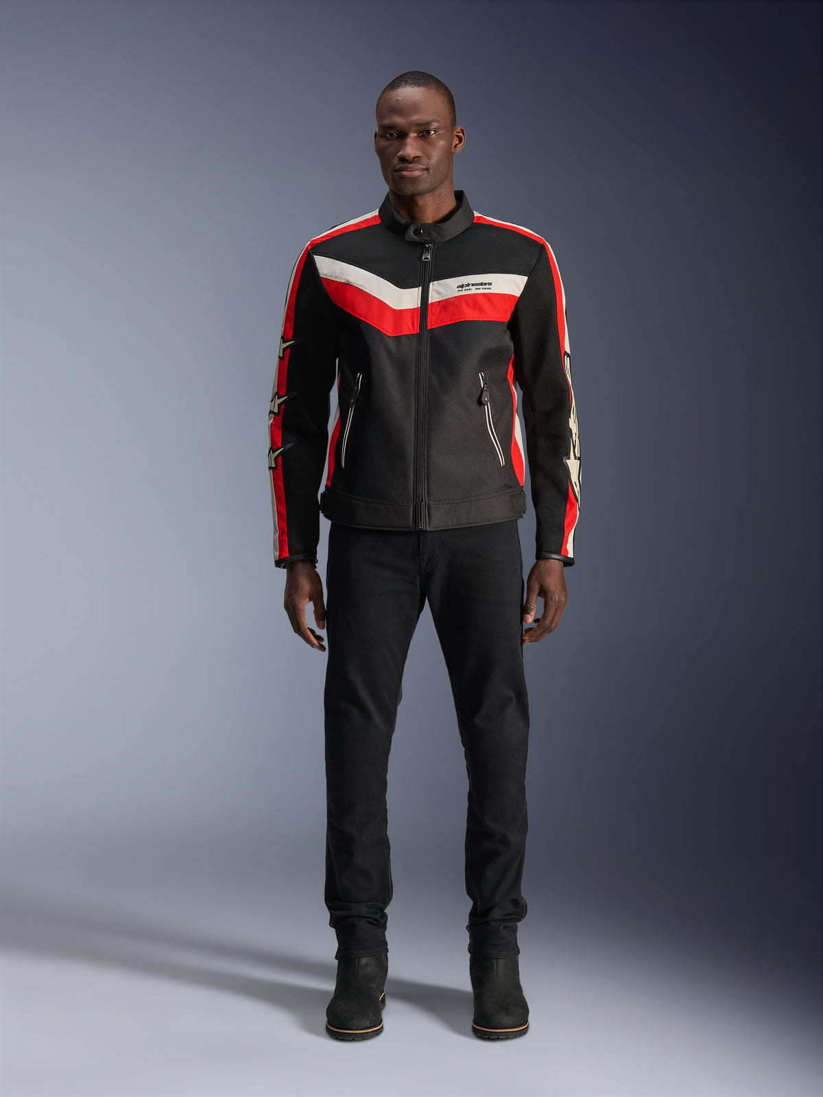 Alpinestars T-Dyno Air-jacka, motorcykeljacka i textil, svart, stark röd och ecru, bärs av en modell med svarta byxor och stövlar, vintageinspirerad design med färgade ränder på bröstet, stjärnlogotyper på ärmarna och ventilerande meshkonstruktion för stadskörning.