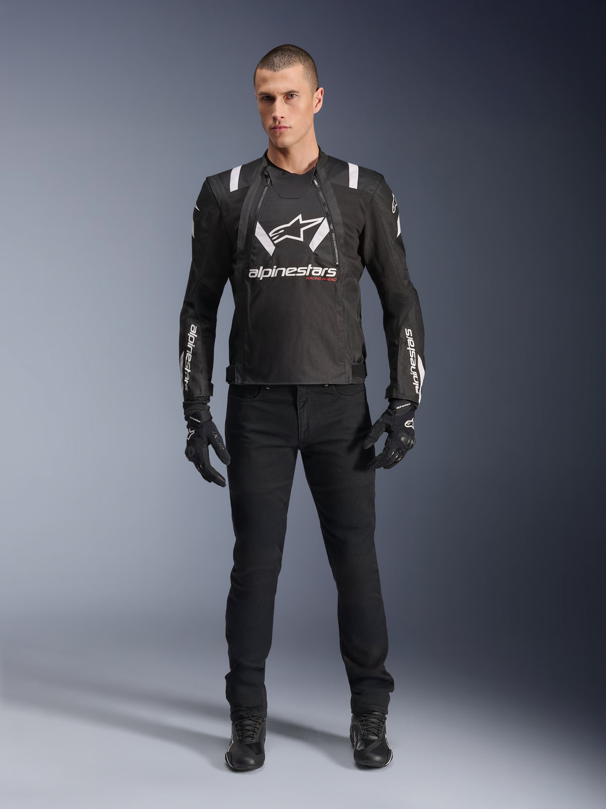 Alpinestars T-Stunt Air-jacka, svart och vit motorcykeljacka, bärs med svarta körbyxor, handskar och stövlar, med framträdande vit Alpinestars-logotyp på bröstet och ärmarna, sportig textildesign med meshpaneler för ventilation