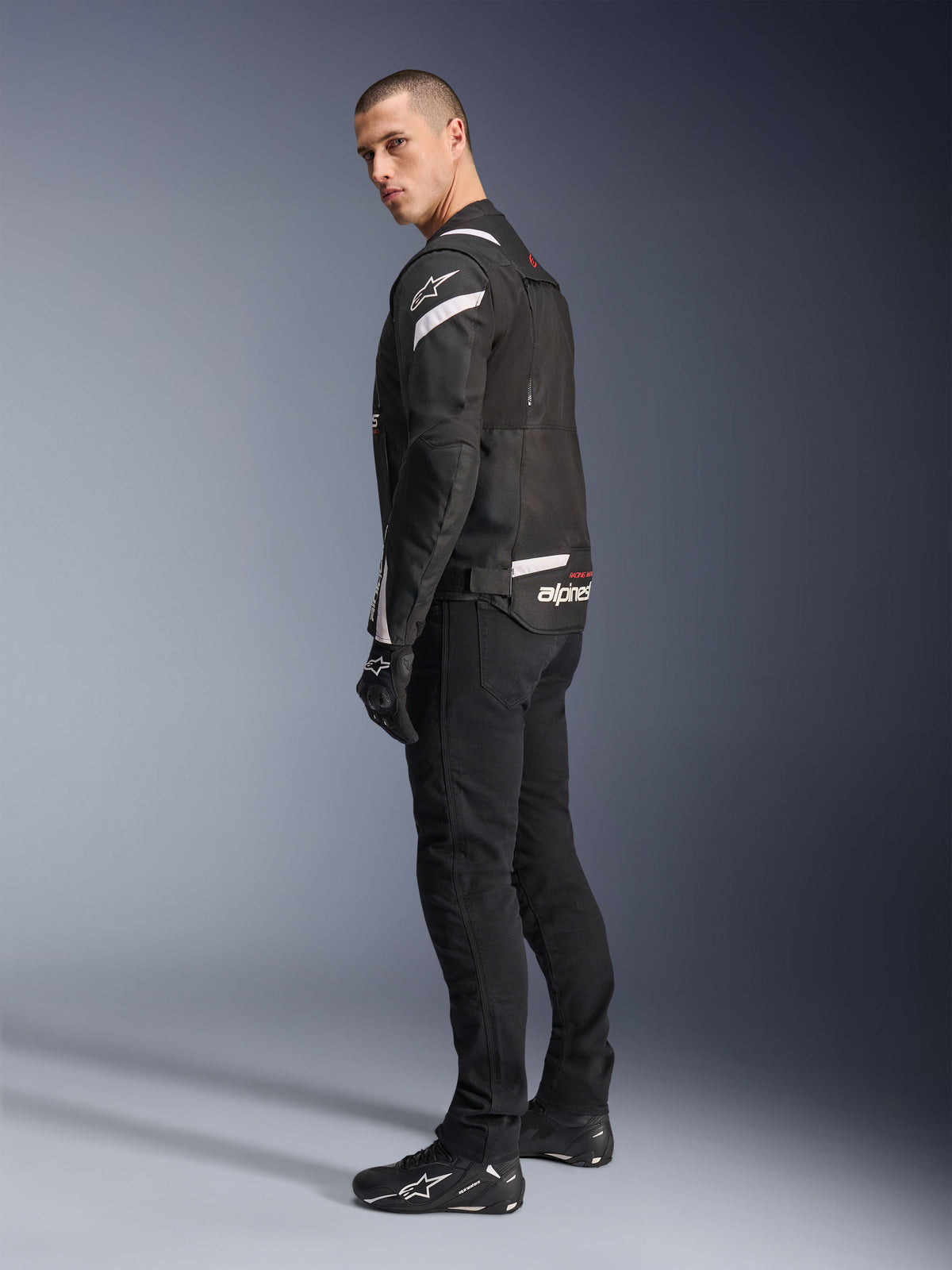 Alpinestars T-Stunt Air Jacket, svart och vit, modulär motorcykeljacka med tech-meshpaneler, visas i profil bakifrån på en modell, bärs med svarta mc-byxor och skor, med vita detaljer och logotyper på axlar och rygg