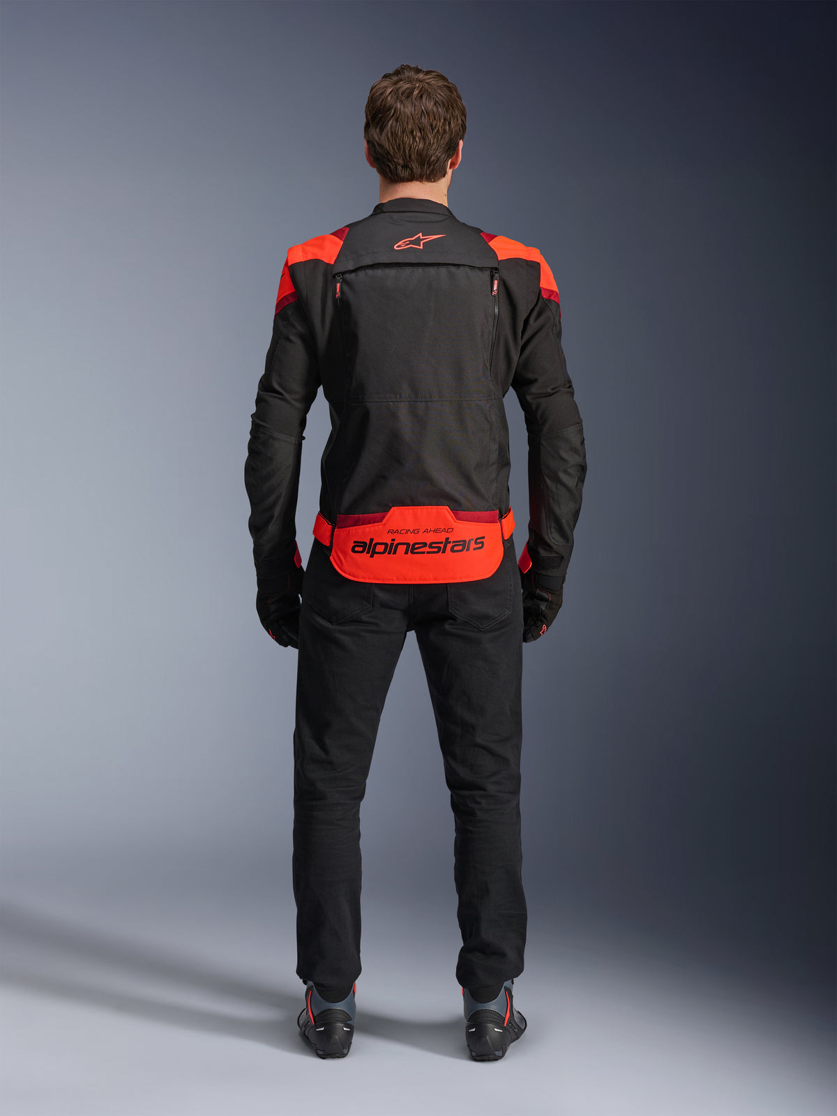 Alpinestars T-Stunt Air Jacket, Black with Red Fluo and Dark Red accents, rear view on model, paired with black riding pants and boots, featuring bold Alpinestars branding on the lower back panel, modular design with mesh construction for ventilation.