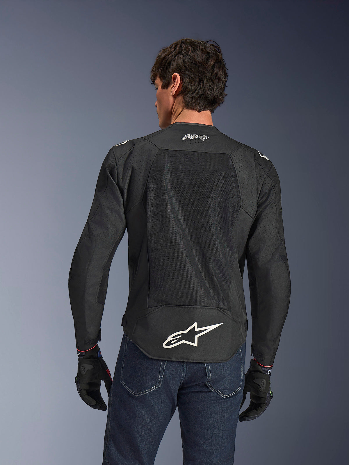 Alpinestars FQ20 T-SPS Air V2 Monster Jacket, Black, Rear view featuring large ventilation mesh panels and white Alpinestars logo on the lower back, worn with blue jeans and black motorcycle gloves, urban sport riding style.
