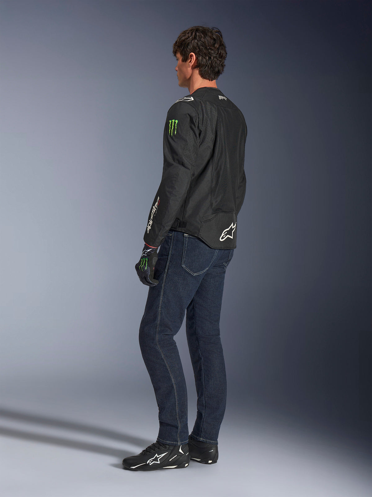 Alpinestars FQ20 T-SPS Air V2 Monster Jacket, black textile motorcycle jacket with green Monster Energy logos on the sleeves and white Alpinestars branding, worn by a model from a rear three-quarter view with dark blue jeans and black riding shoes, urban sport style.