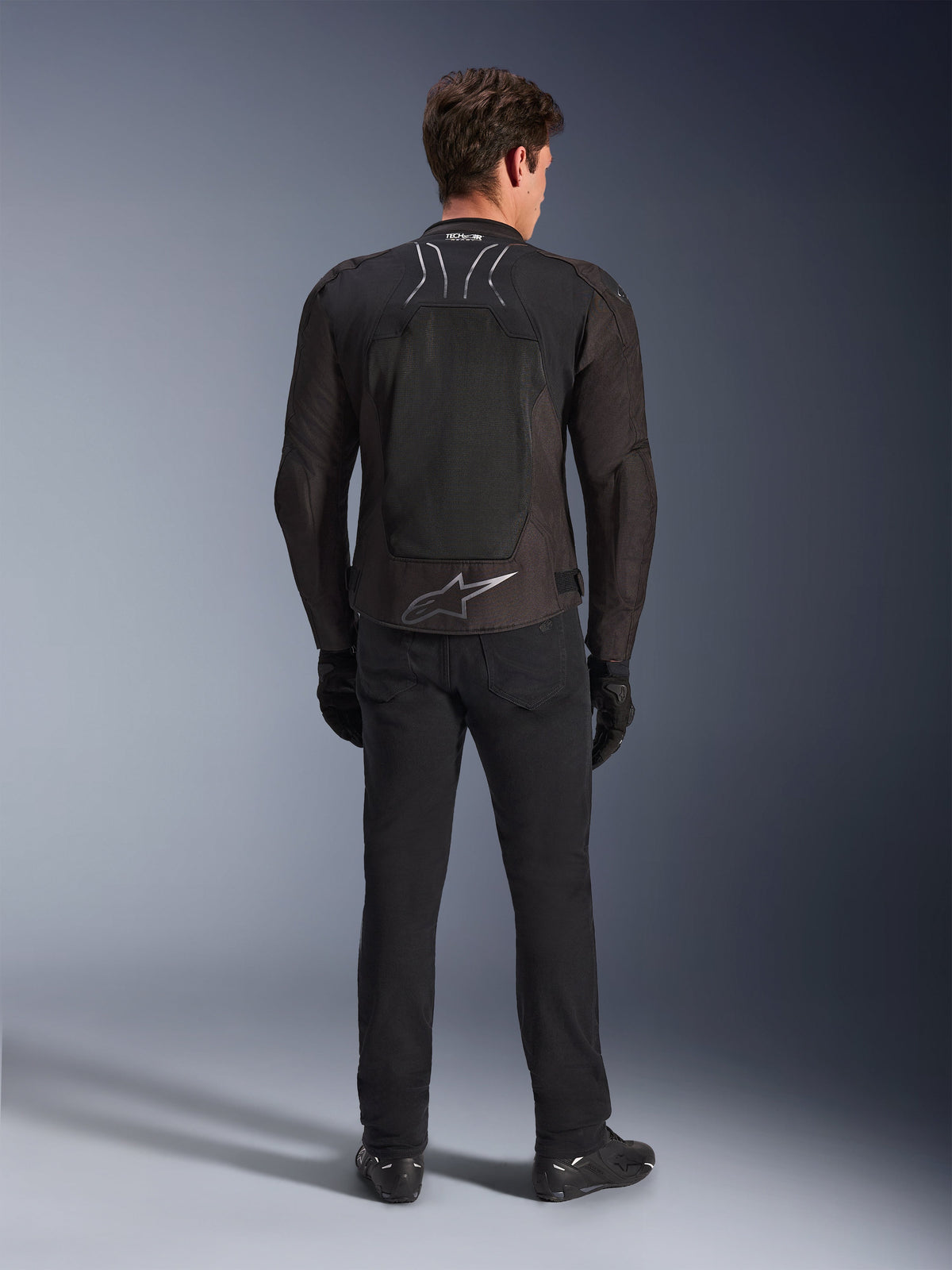 Alpinestars SMX Air-jacka, Black Anthracite, ryggvy på manlig modell, med en stor silverfärgad Alpinestars-logotyp på nedre delen av ryggen och meshpaneler för högt luftflöde, bärs med svarta mc-byxor, handskar och stövlar, sportig motorcykelutrustning