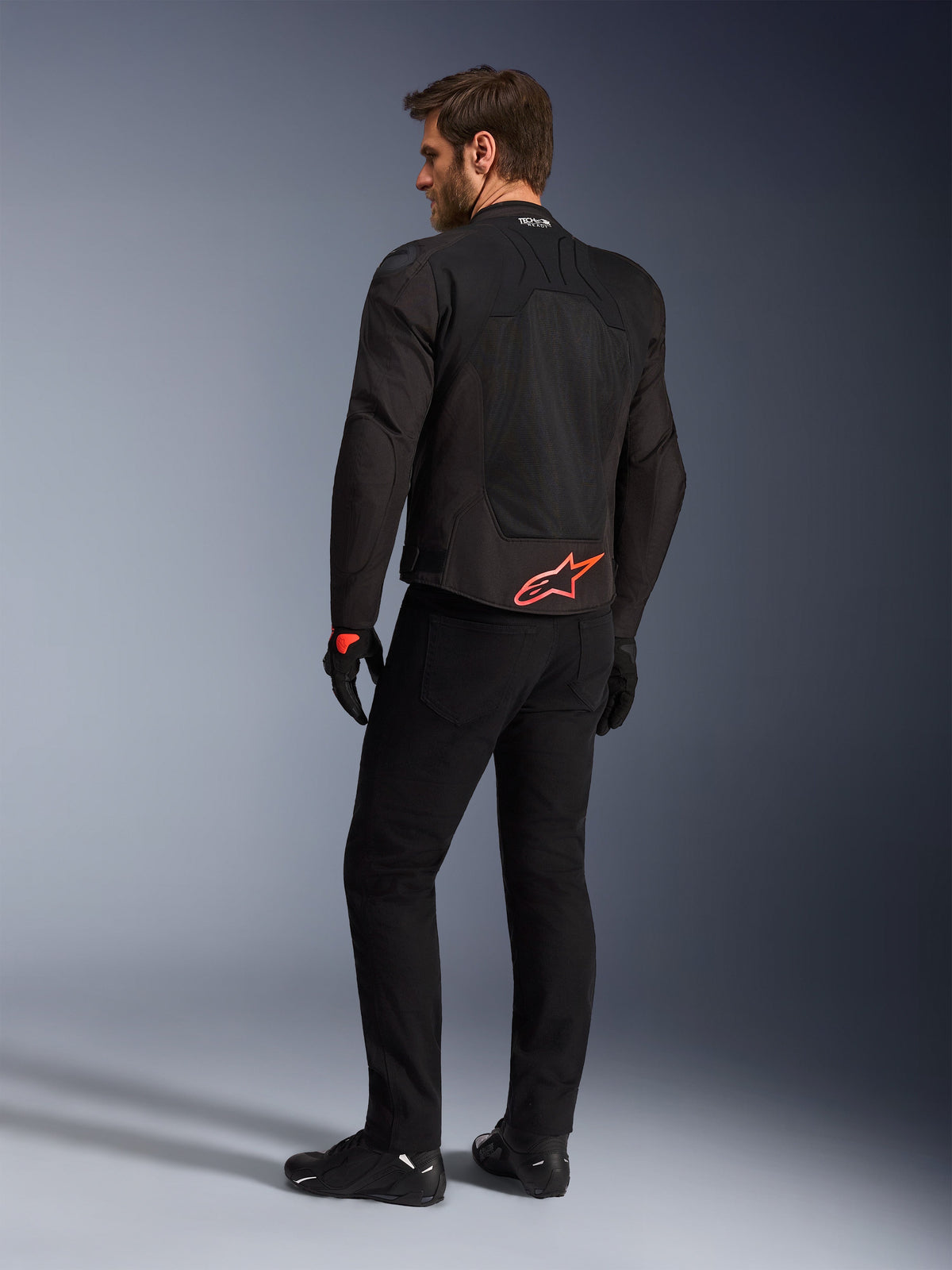 Bakifrån vy av en modell som bär Alpinestars SMX Air Jacket i svart, med en klaröd Alpinestars-logotyp på nedre delen av ryggen, 'Tech-Air Ready'-märkning på kragen och tekniska mesh-paneler för ventilation, matchad med svarta körbyxor och handskar.