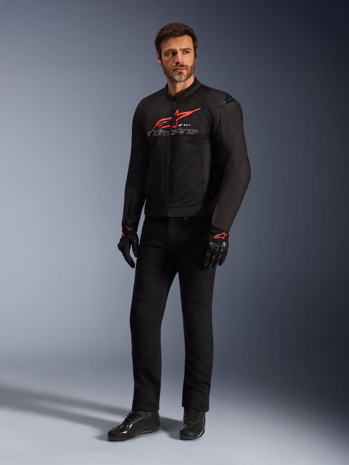Manlig modell som bär Alpinestars SMX Air-jacka i svart, klarrött och vitt, stylad med svarta tekniska byxor, svarta och röda handskar och svarta mc-skor, med en framträdande röd logotyp på bröstet och vit branding, helkroppsbild i studiomiljö för sportmotorcykelutrustning