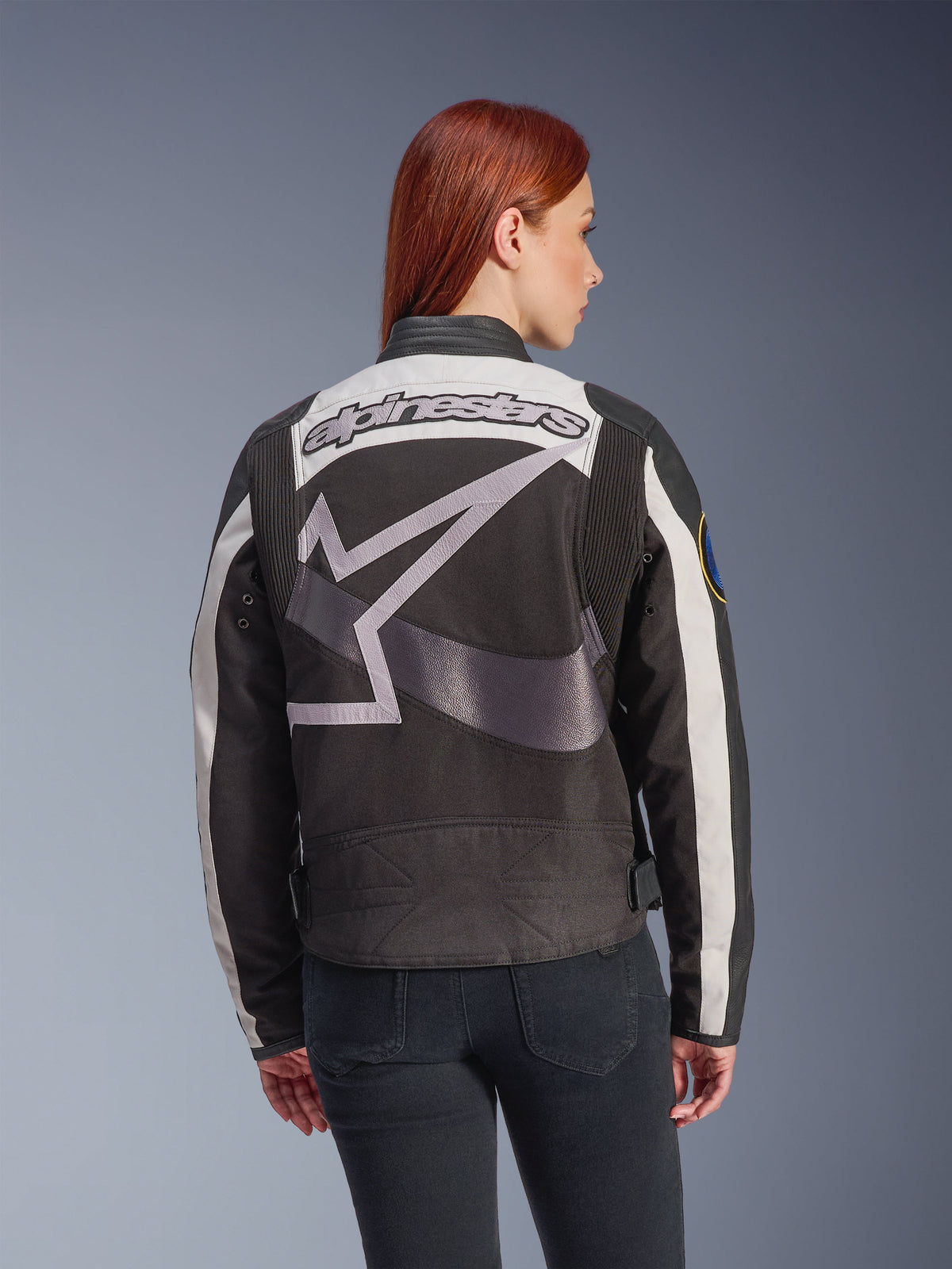 Alpinestars Idles damjacka, svart och off-white, baksida med en stor grå stjärnlogotyp och Alpinestars ordmärke, bärs av en rödhårig modell, vintageinspirerad urban motorcykeljacka med dragspelspaneler.
