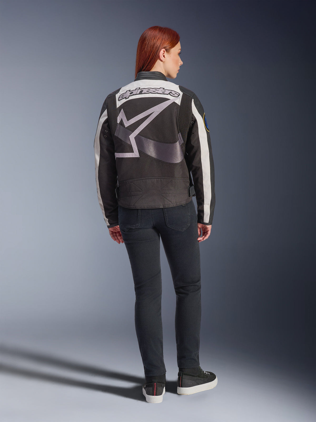 Alpinestars Idles damjacka, urban motorcykeljacka, svart och off-white, ryggvy med en stor grå Alpinestars-logotyp och ordmärke, bärs med svarta slim-fit jeans och sneakers, vintageinspirerad design för stadskörning