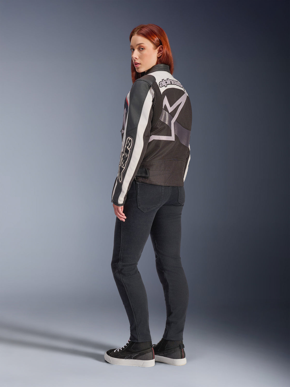 Kvinna som bär Alpinestars Idles-jacka, ryggvy, Off White Flame Red och svart, med en stor stjärnlogotyp på ryggen och Alpinestars-ordmärke på ärmen, kombinerat med svarta slim-fit-byxor och sneakers, urban motorcykelutrustning för damer