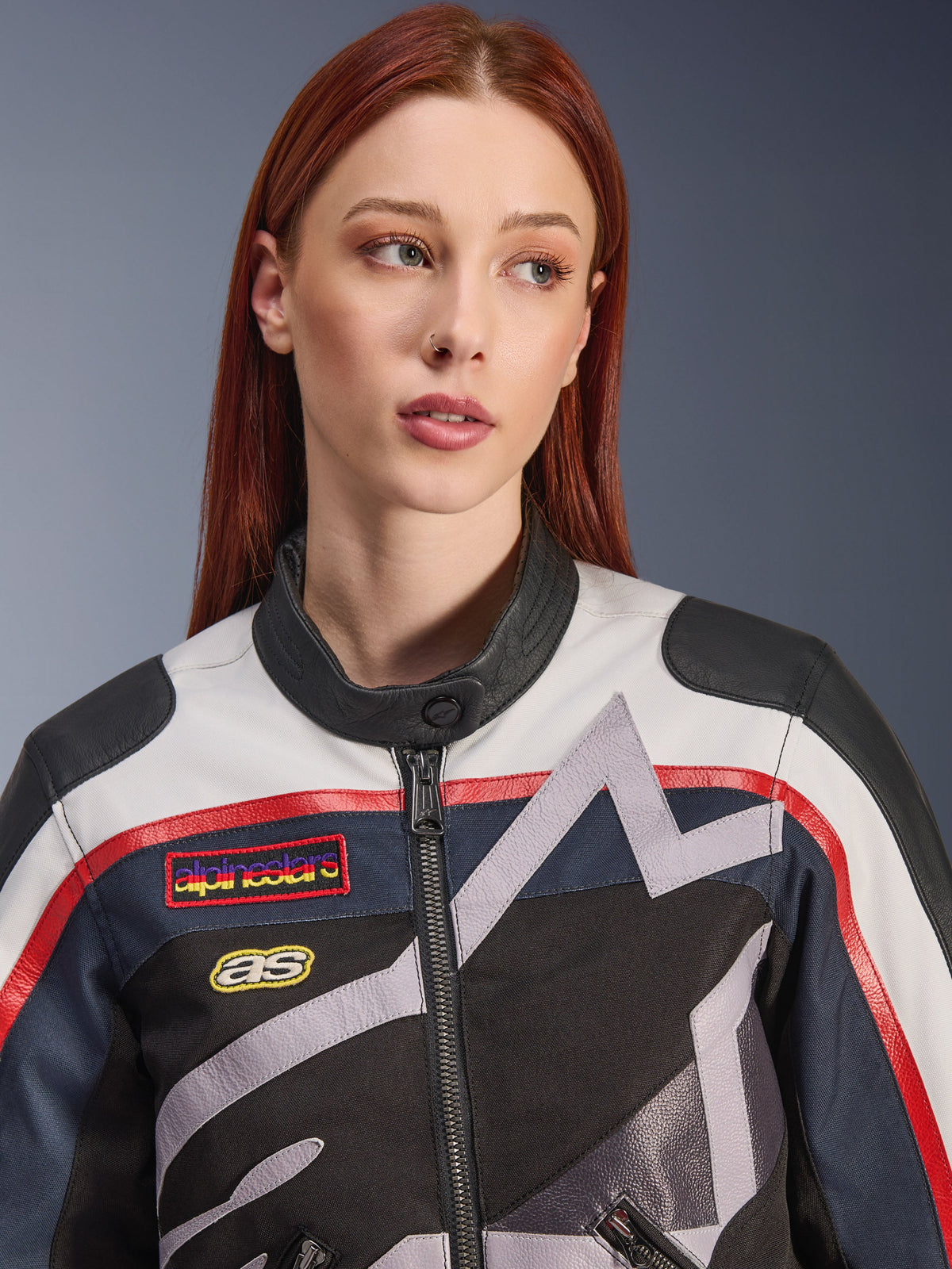 Alpinestars Idles damjacka, närbild på en kvinnlig modell med rött hår, färgställningen Off White Flame Red och Black, vintageinspirerad racingdesign med logotyp-patchar och grå geometrisk stjärngrafik, urban mc-utrustning.
