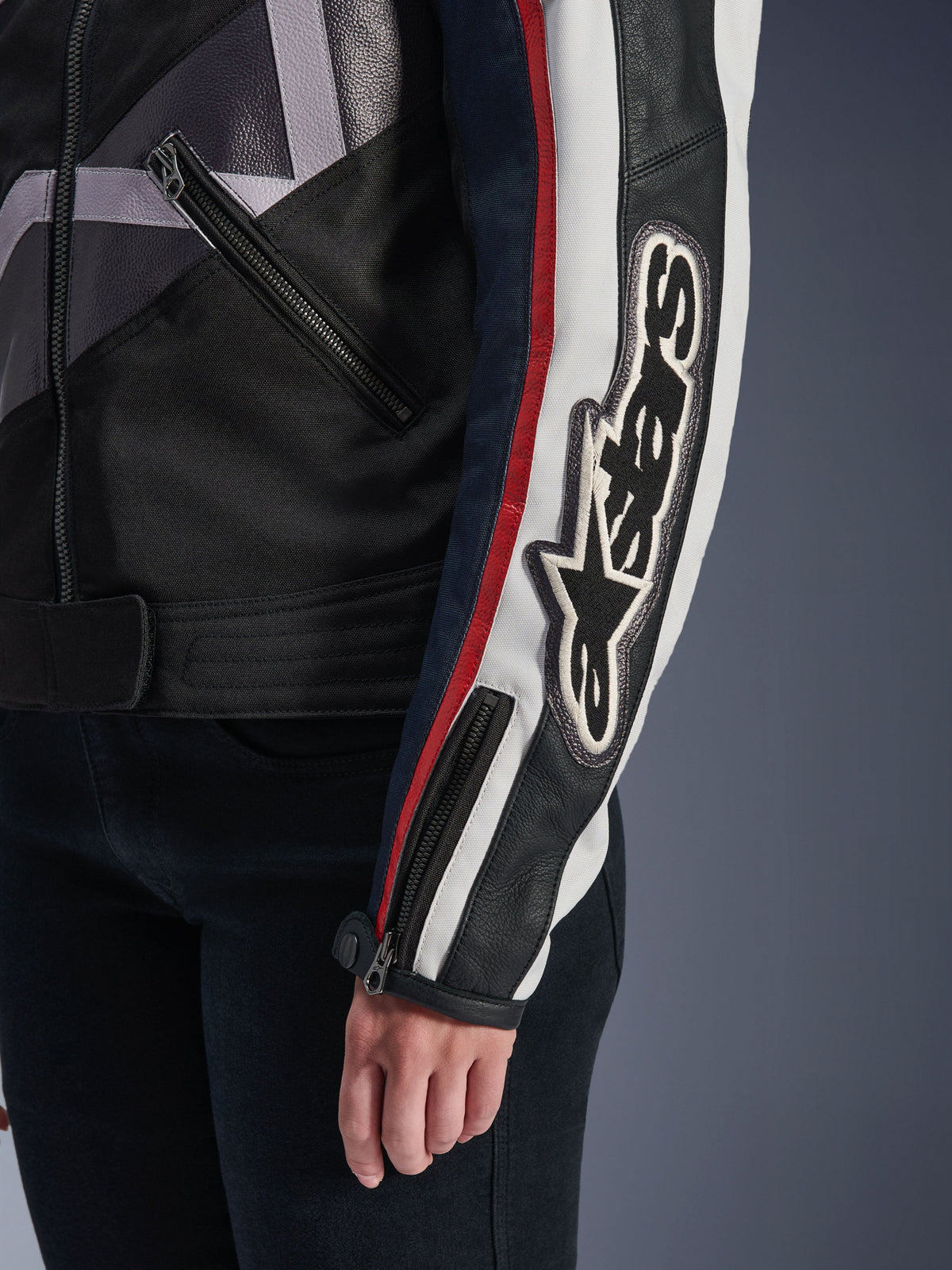 Alpinestars Idles damjacka, urban motorcykeljacka, Off White Flame Red Black, närbild på ärmen med en stor broderad Alpinestars-logotyp, röda och vita racingränder, konstruktion i svart textil och läder samt detaljer med metalldragkedjor.