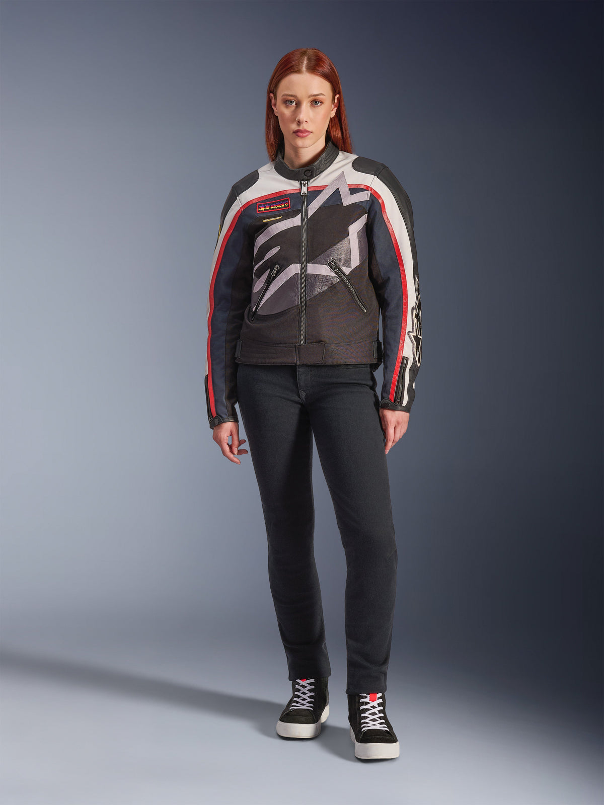 Kvinnlig modell bär Alpinestars Idles Jacket, motorcykeljacka för dam, Off White, Flame Red och svart, med en stor omslutande stjärnlogotyp och vintageinspirerade detaljer, kombinerad med mörka slim-fit körbyxor och svarta high-top sneakers, urban modern motorcykelutrustning