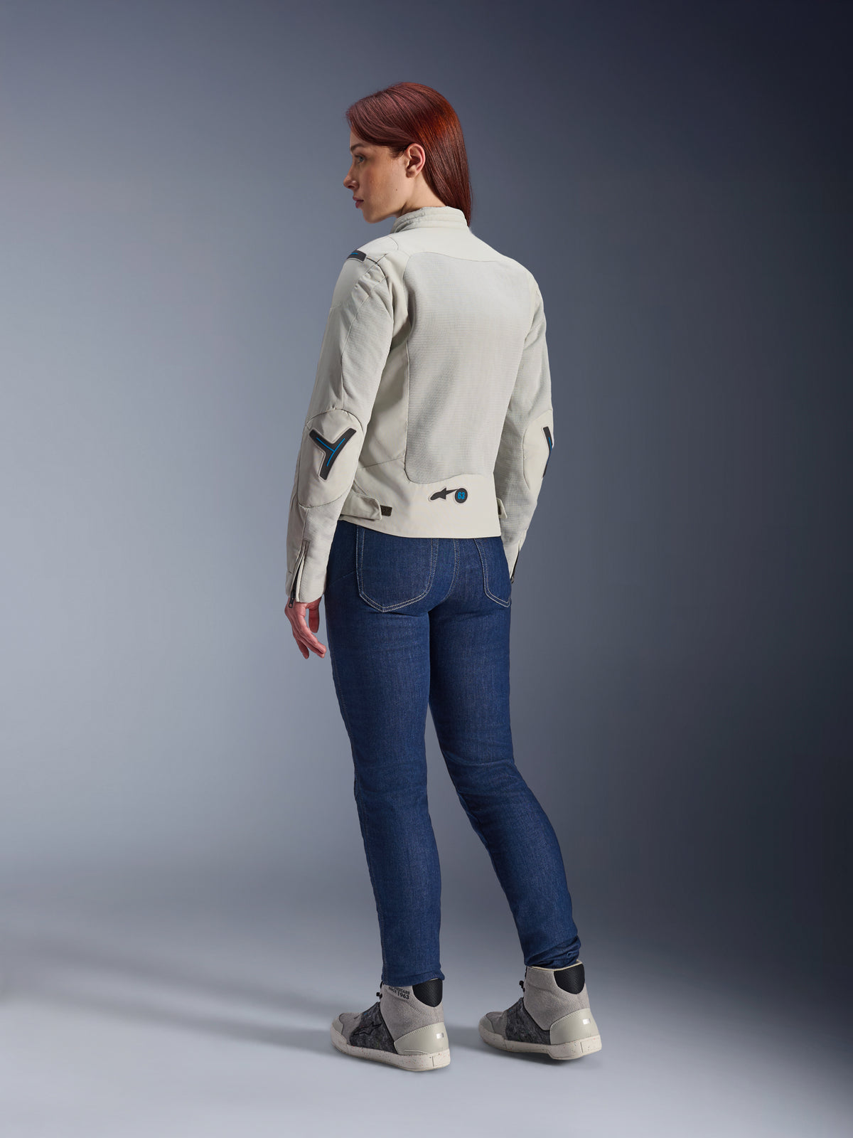 Kvinna i Alpinestars Stella Circal-1 Air Jacket i Metal Gray och Methyl Blue, vy bakifrån som visar meshpaneler för ventilation och neonblå detaljer, kombinerat med mörkblå mc-jeans och grå sneakers, urban livsstilsbild för motorcykel.