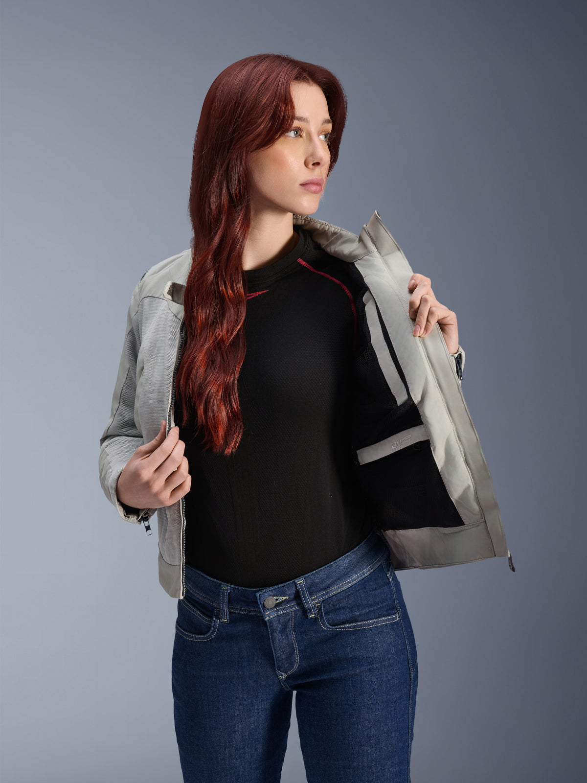 Alpinestars Stella Circal-1 Air-jacka i Metal Gray, kvinnlig modell som öppnar jackan för att visa det inre meshfodret och fickan, bärs med blå jeans och en svart topp, modern urban motorcykelutrustning.