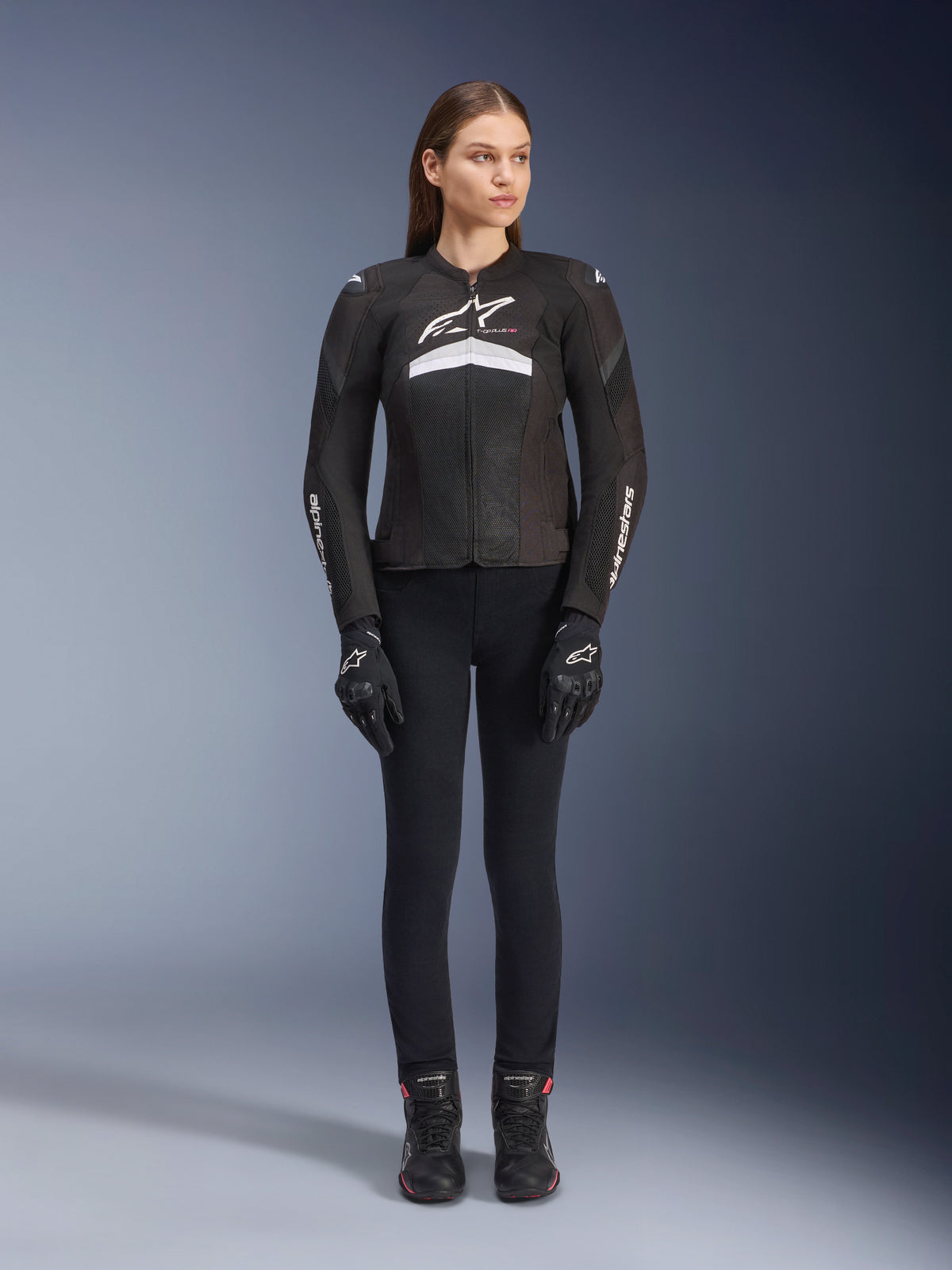 Kvinnlig förare i Stella T-GP Plus R V4 Airflow-jacka, svart och vit, matchad med svarta körbyxor, handskar och stövlar, med meshpaneler för ventilation och vit Alpinestars-logotyp på bröstet och ärmarna.